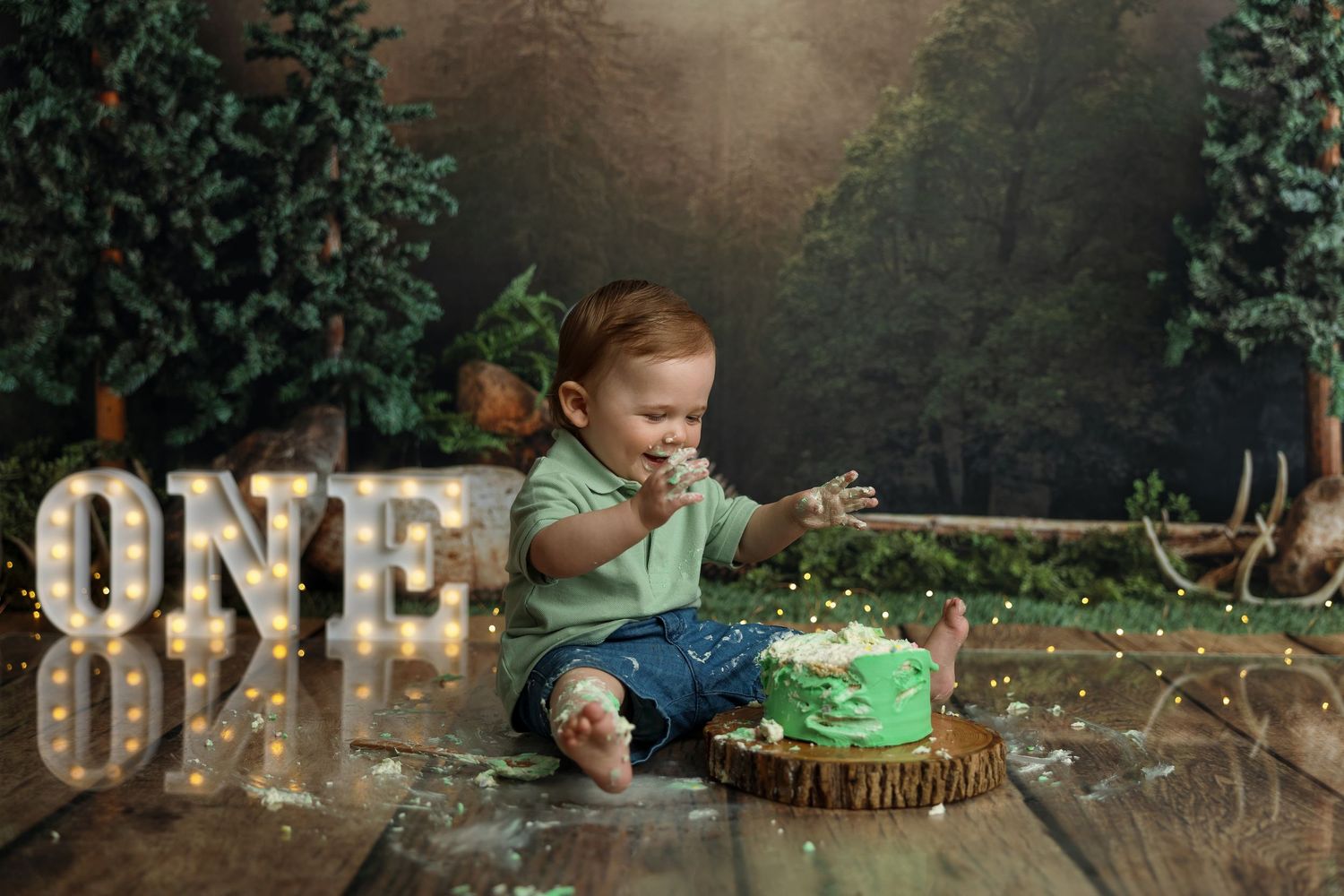Baby's first birthday cake smash photo with green cake, ONE marquee sign, and forest backdrop on wooden floor.