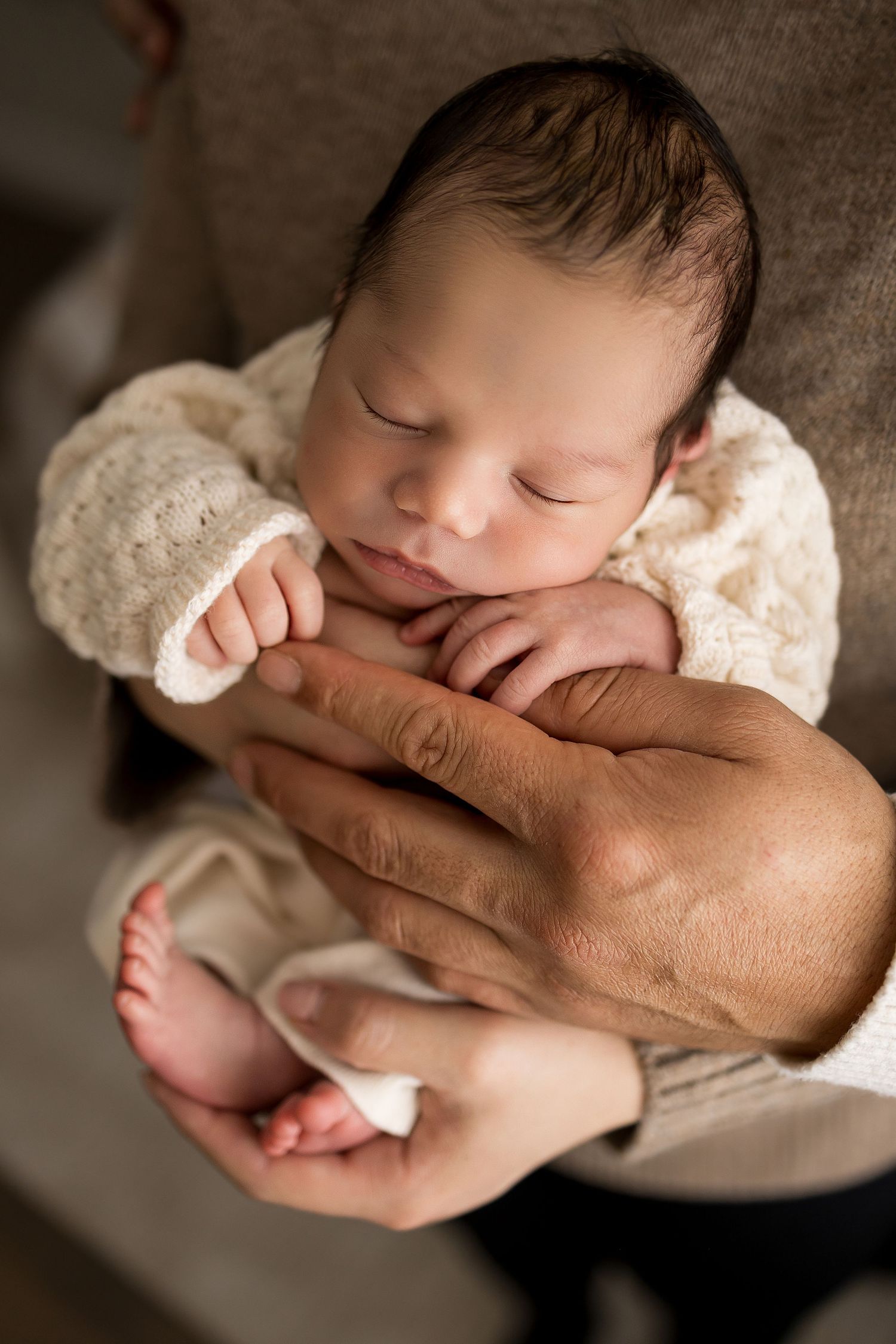 Sleeping newborn baby in white knit outfit cradled gently in adult hands, peaceful and serene newborn photography.
