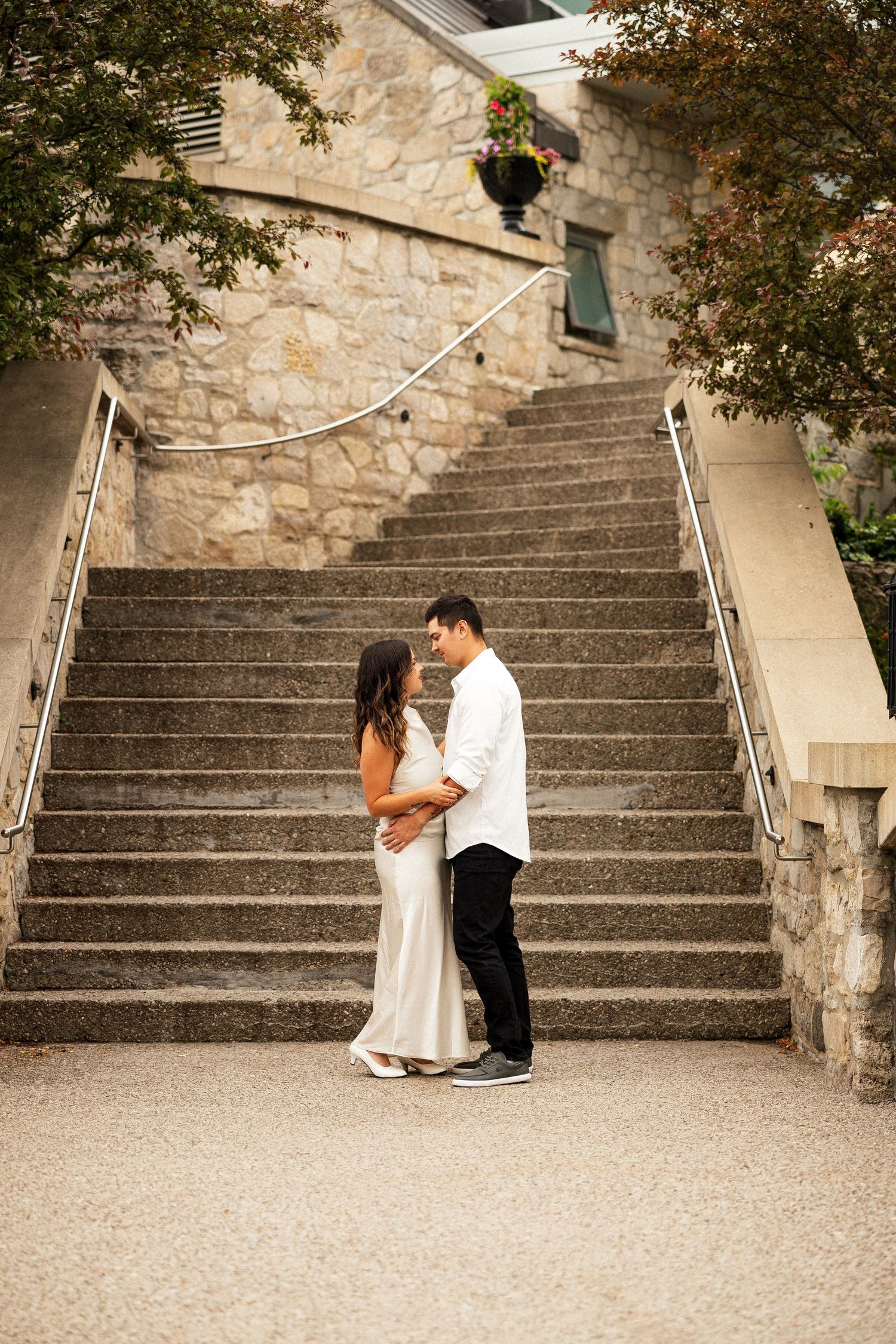 Couple in elegant attire embracing at the base of stone stairs during an outdoor engagement photo session.