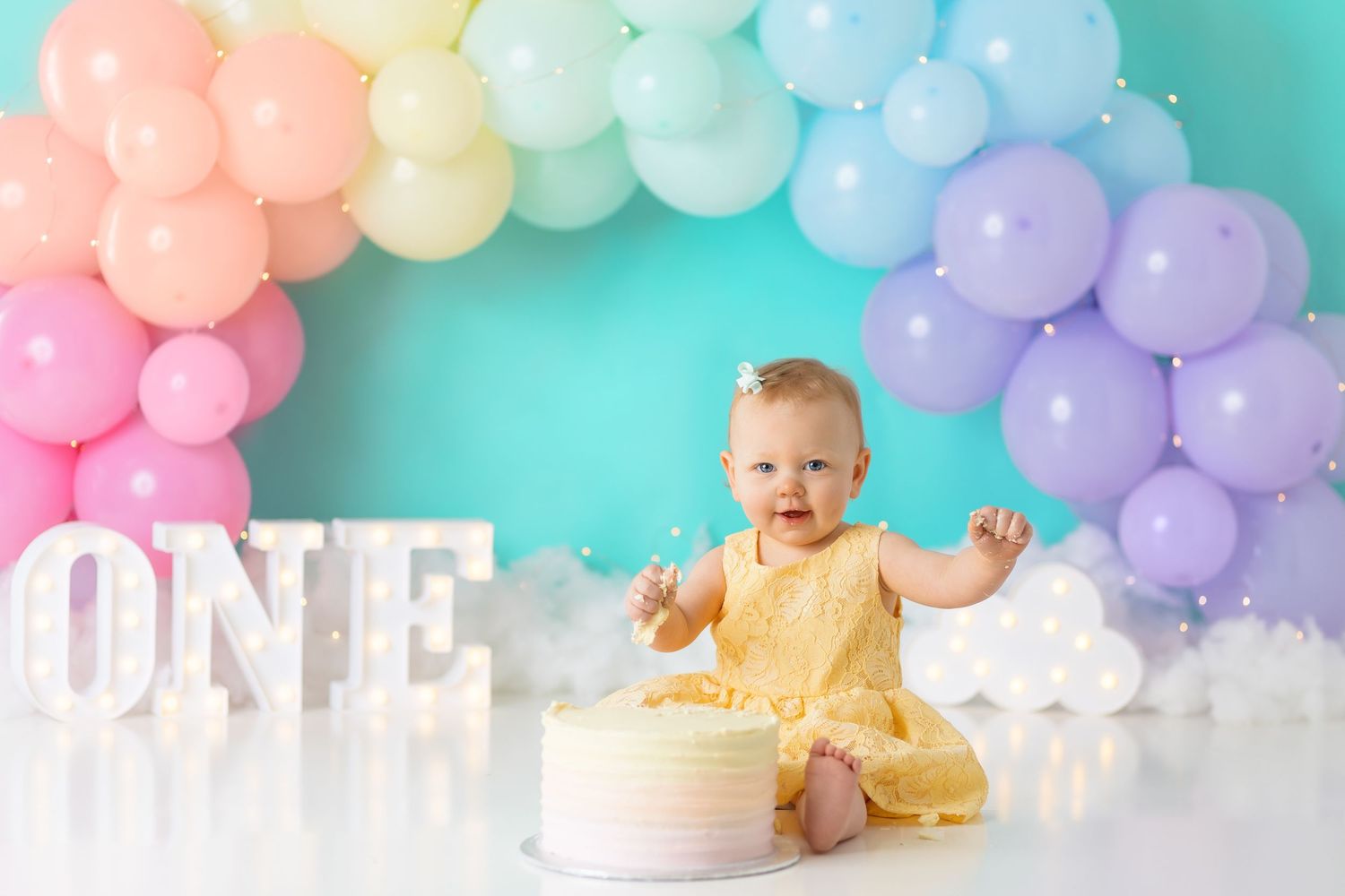 Pastel rainbow balloon arch with white ONE letters and yellow cake for first birthday.
