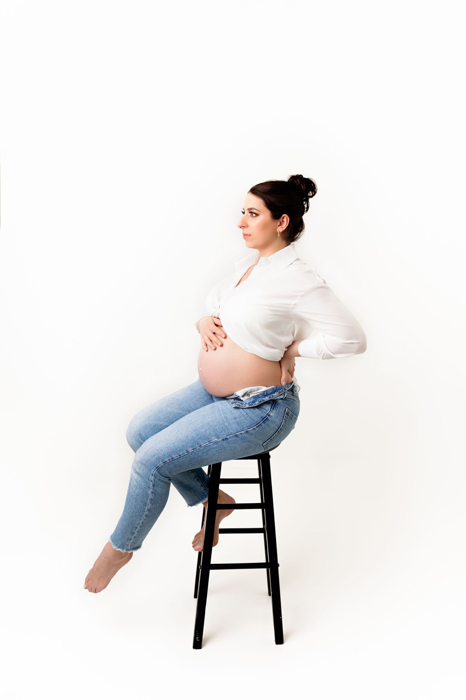 Expectant mother in casual attire sits on black stool during minimalist studio photography session.