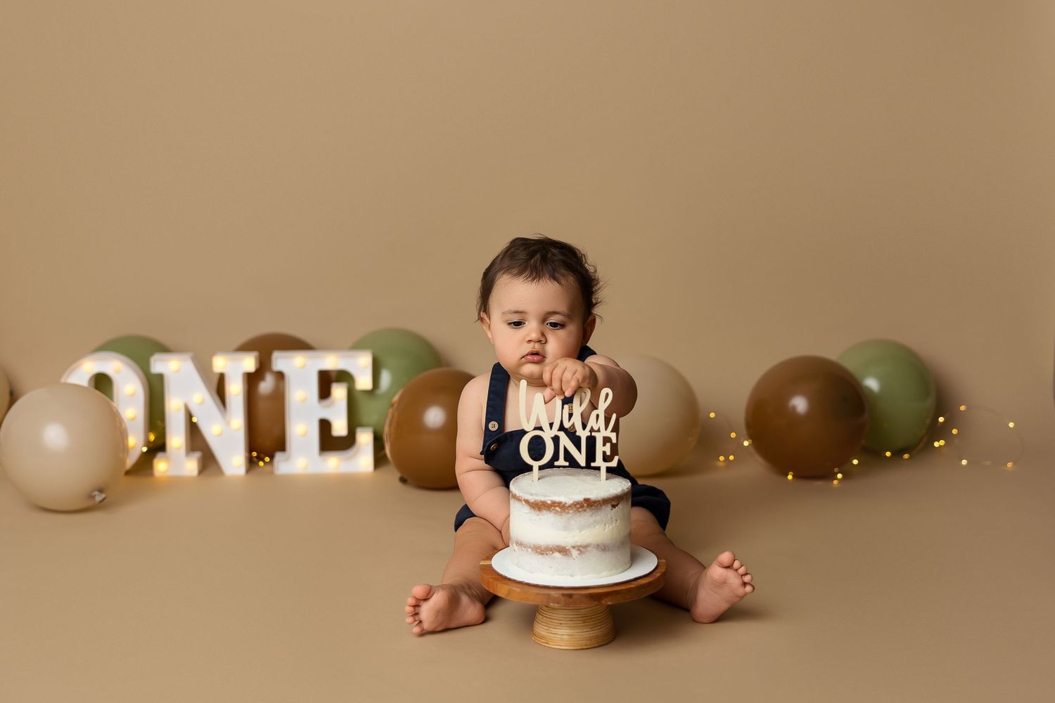 First birthday cake smash photo shoot with white 'ONE' text and brown and green balloons on beige backdrop.