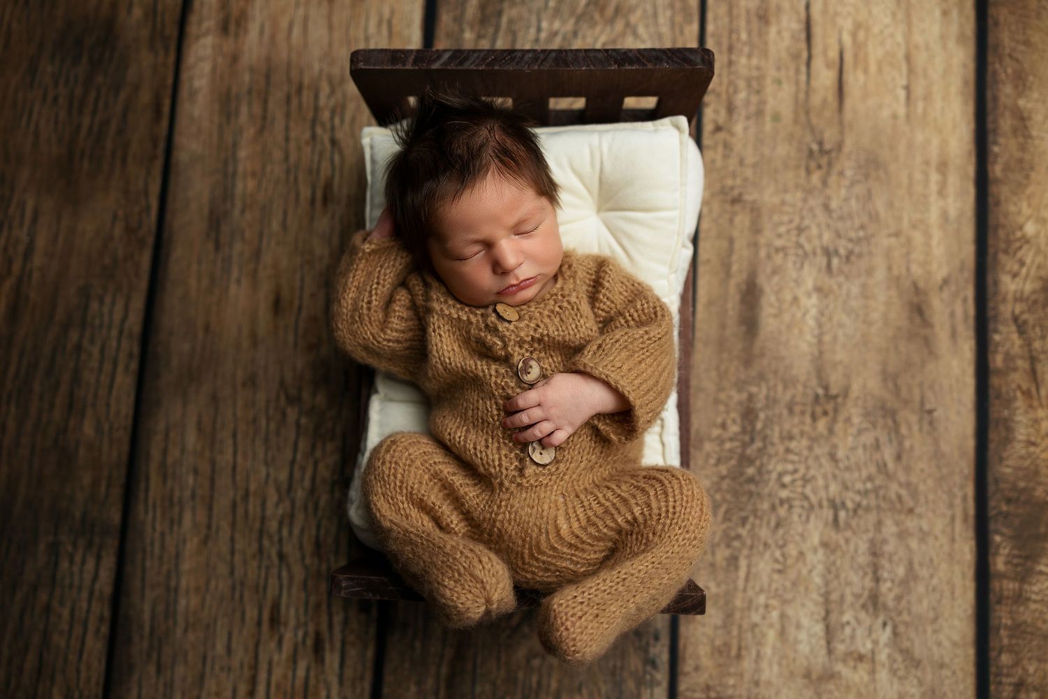 Newborn baby sleeping peacefully in brown knit wrap against rustic wooden backdrop in professional portrait setting.