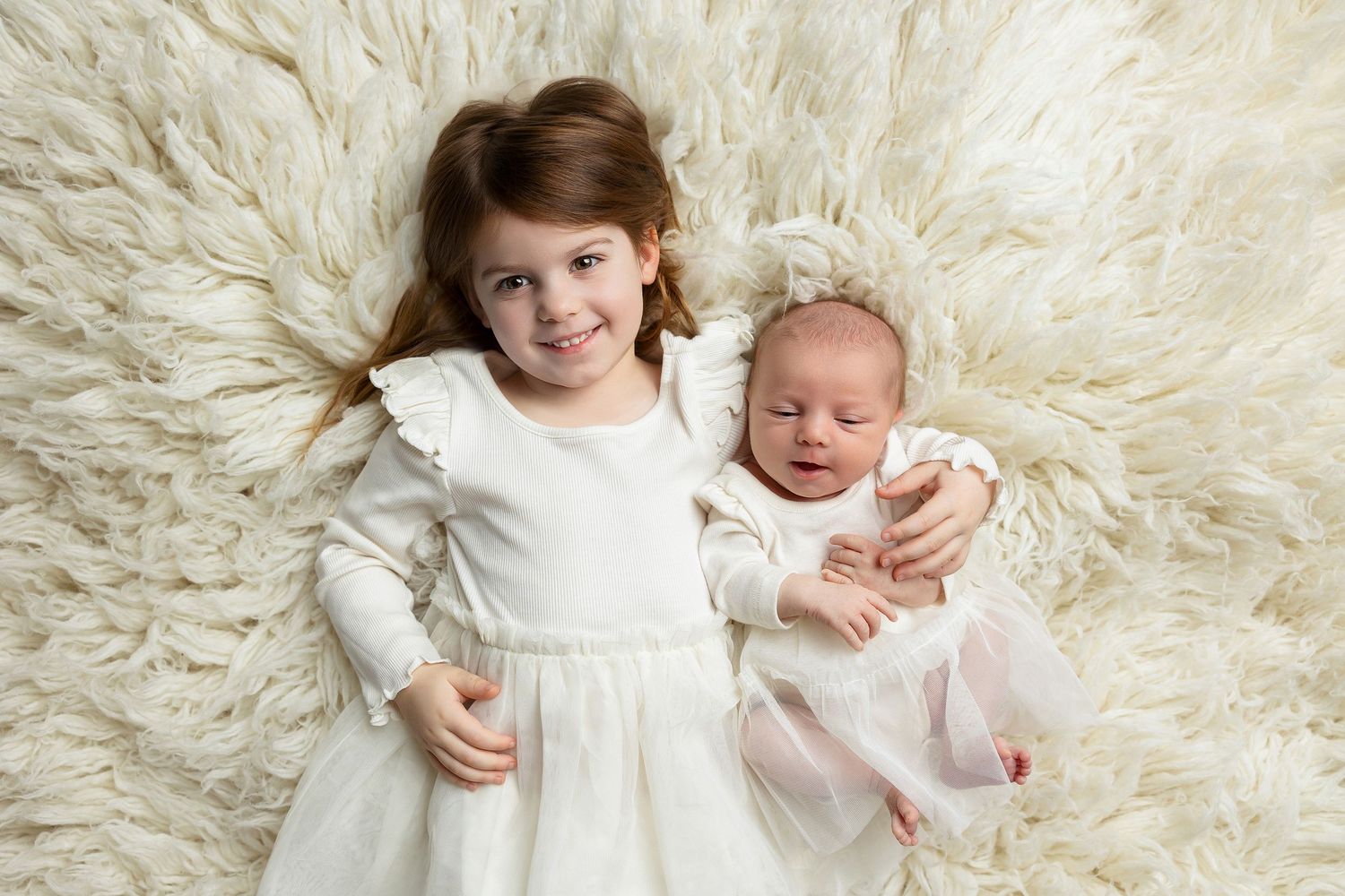 Two children in white dresses lying on a fluffy cream-colored fur background.