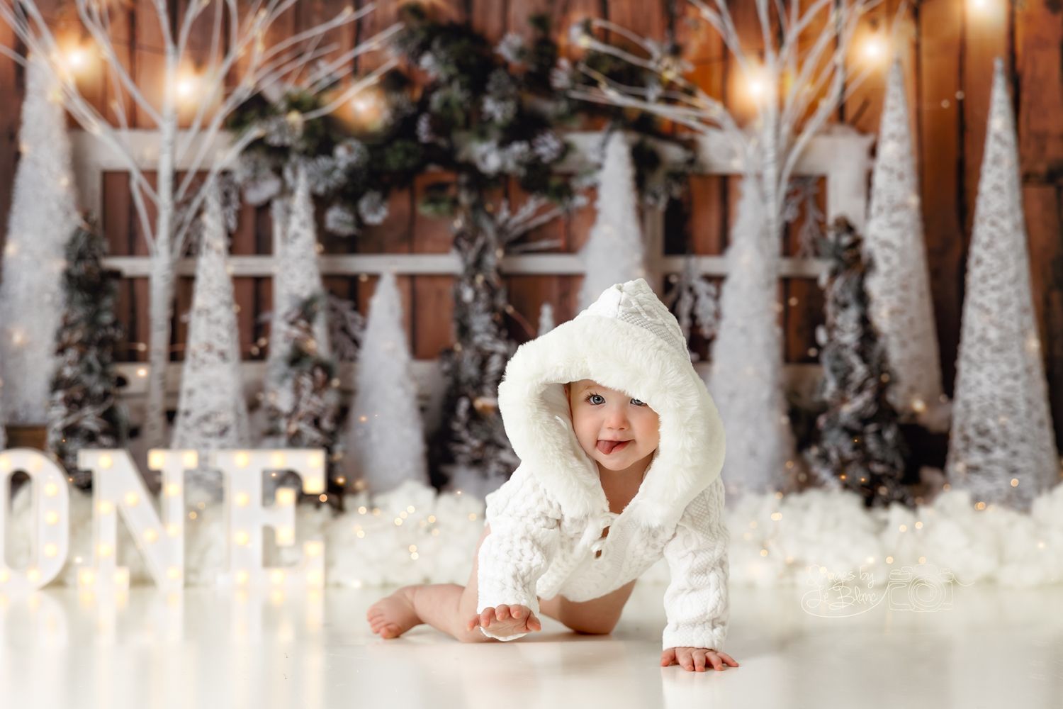 Baby in white winter outfit crawling near illuminated Christmas decorations and snowy trees.