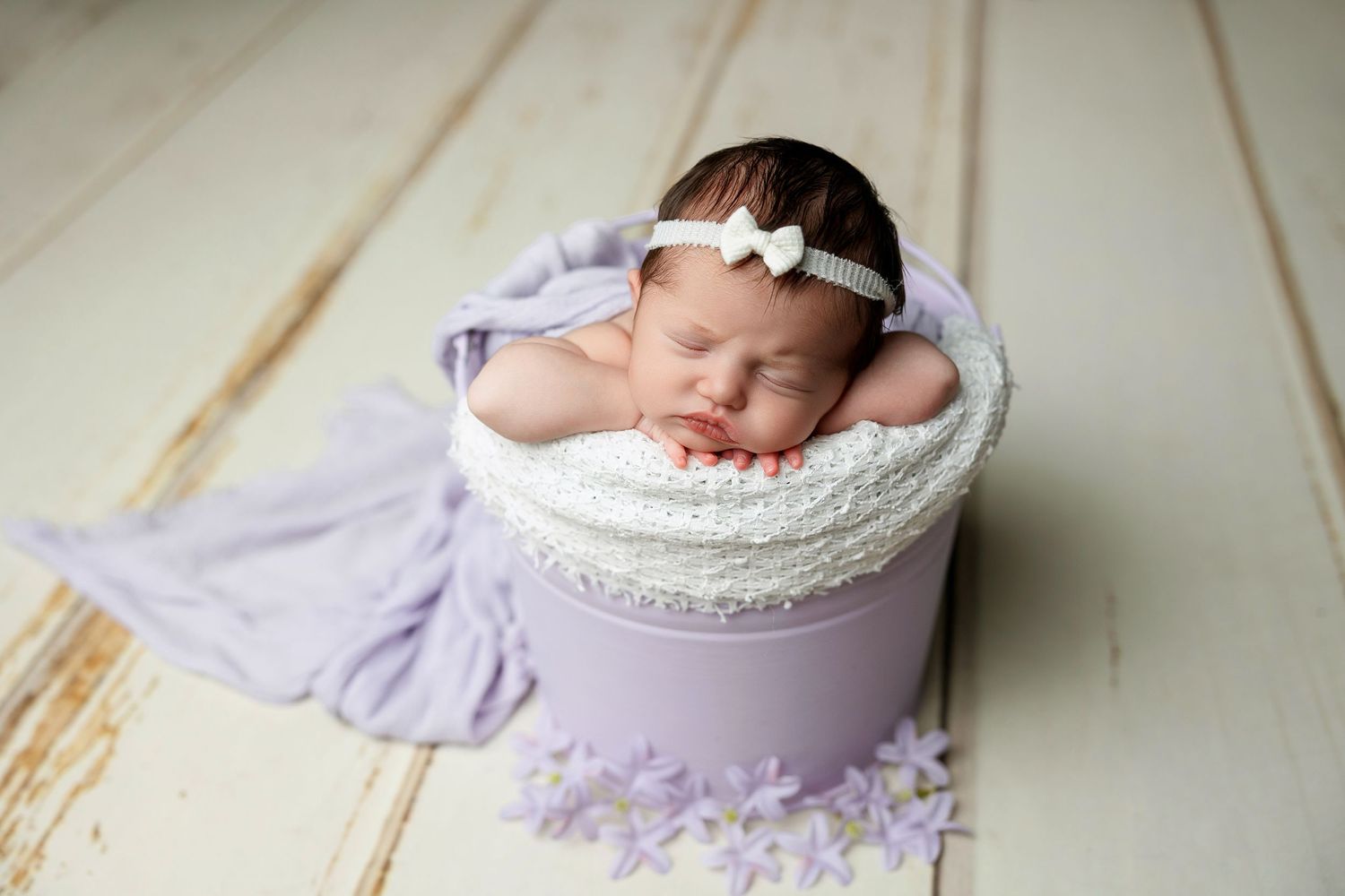 Sleeping newborn with bow headband nestled in purple bucket with lace trim and fabric draping.