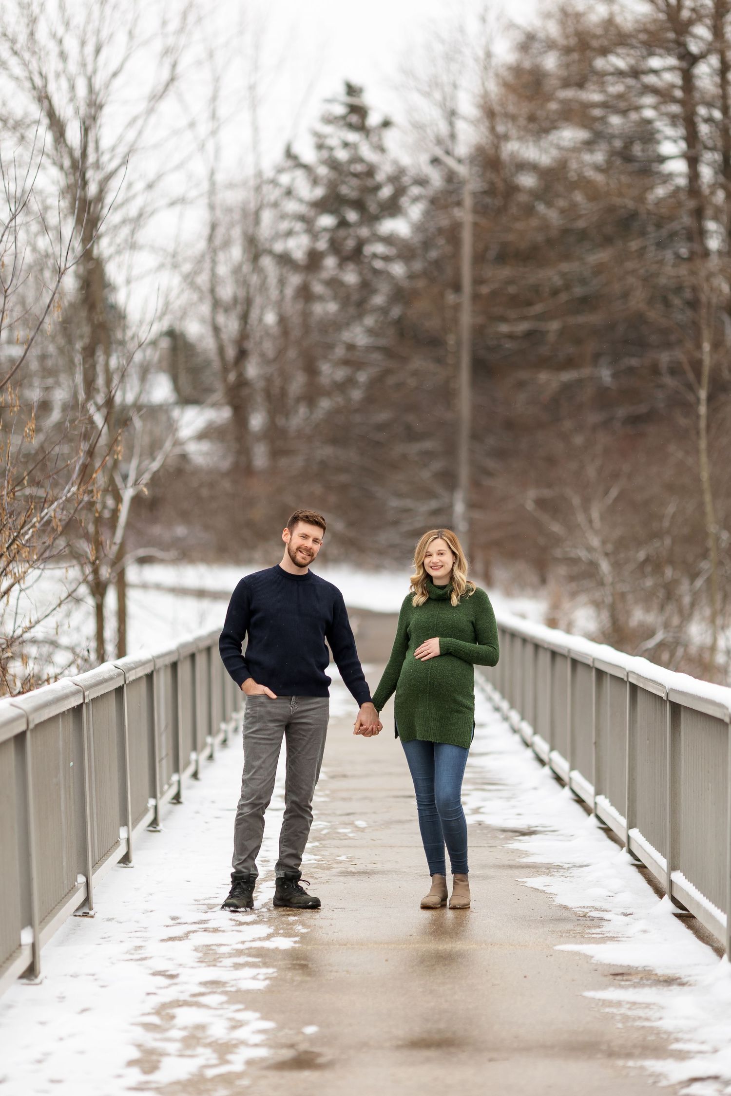 Expectant couple walking hand in hand on snowy bridge in winter.