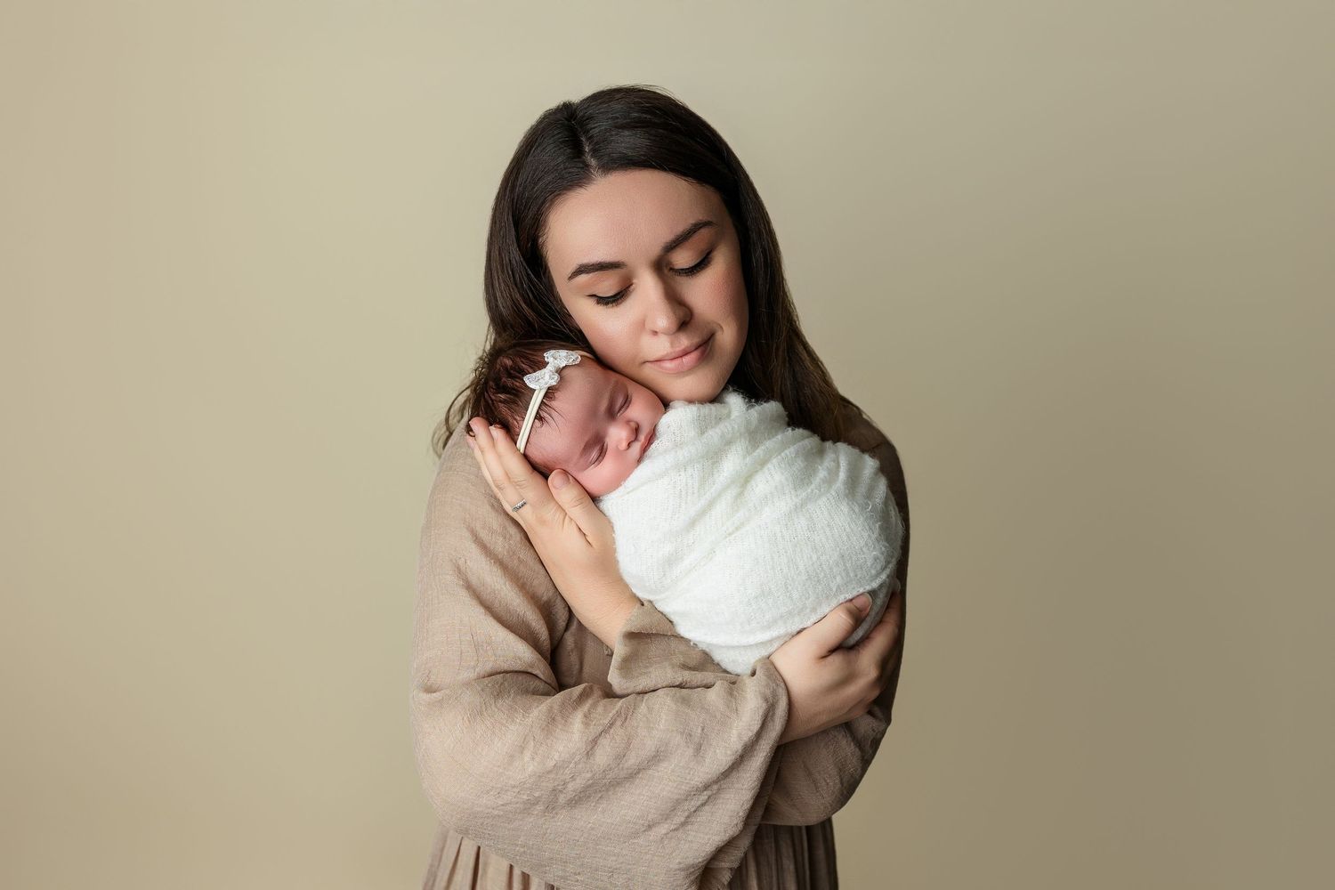 Woman in beige sweater tenderly holding newborn baby wrapped in white blanket against neutral background.