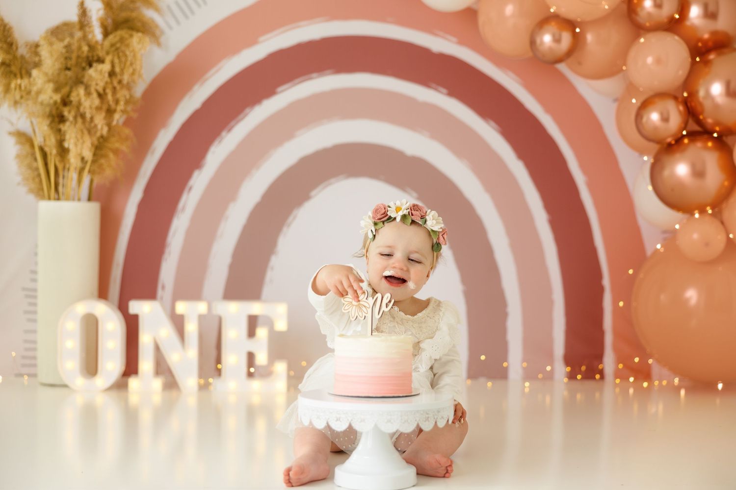 First birthday celebration setup with pink rainbow backdrop, floral crown, white dress and decorative balloons.
