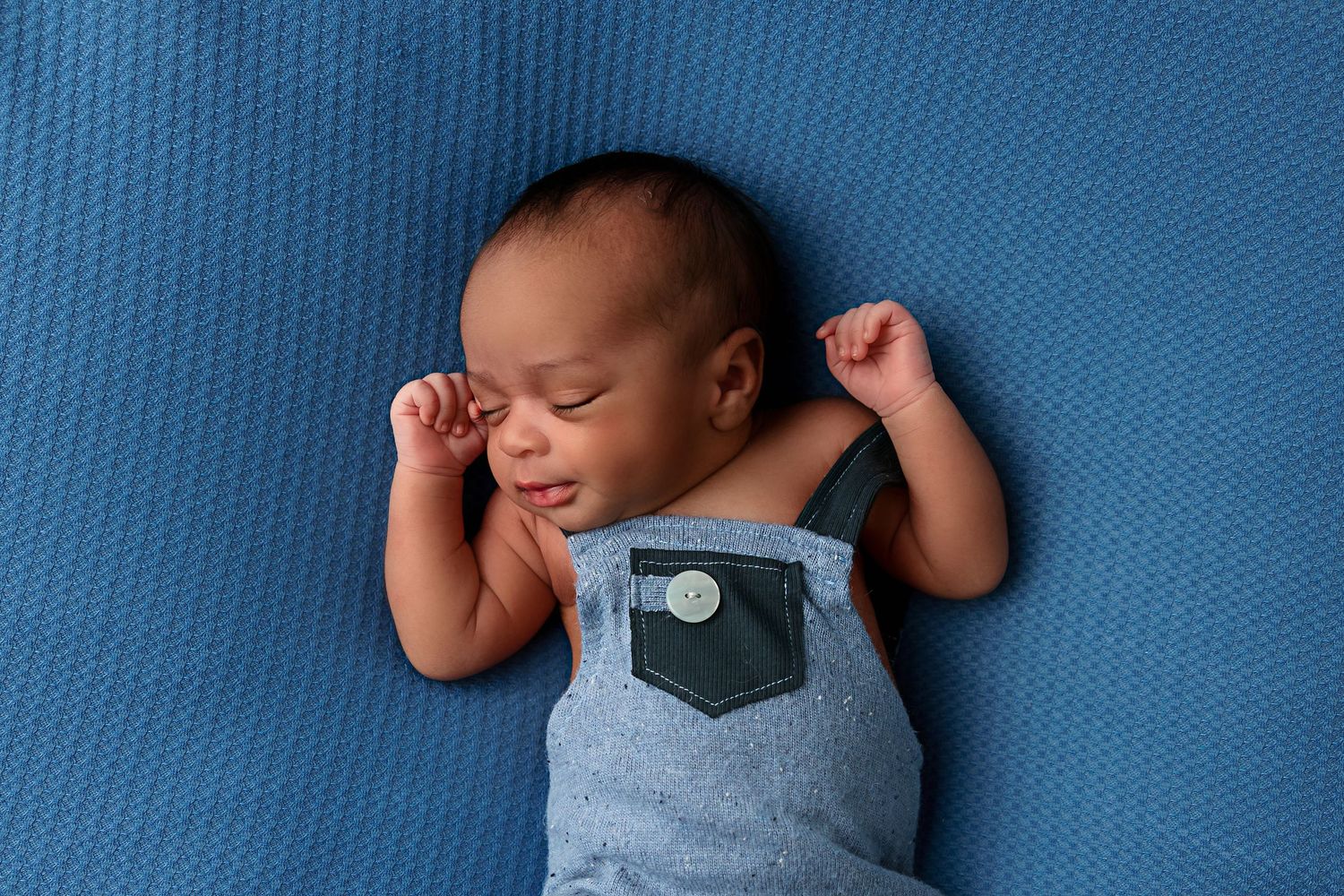 Sleeping baby wearing blue overalls stretches arms overhead on blue backdrop.