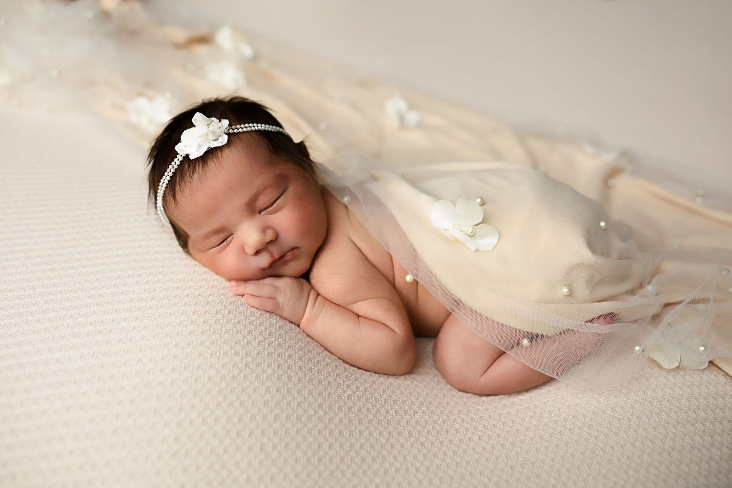 Peaceful sleeping baby with floral headband draped in sheer cream fabric on white backdrop.