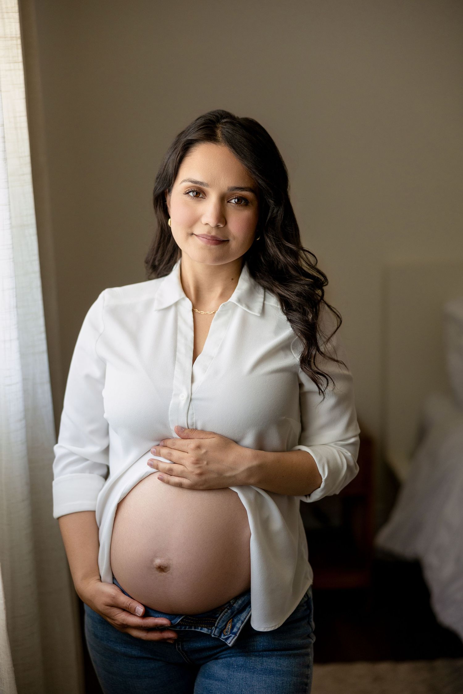 A woman in a white button-down shirt cradles her pregnant belly against a neutral backdrop.