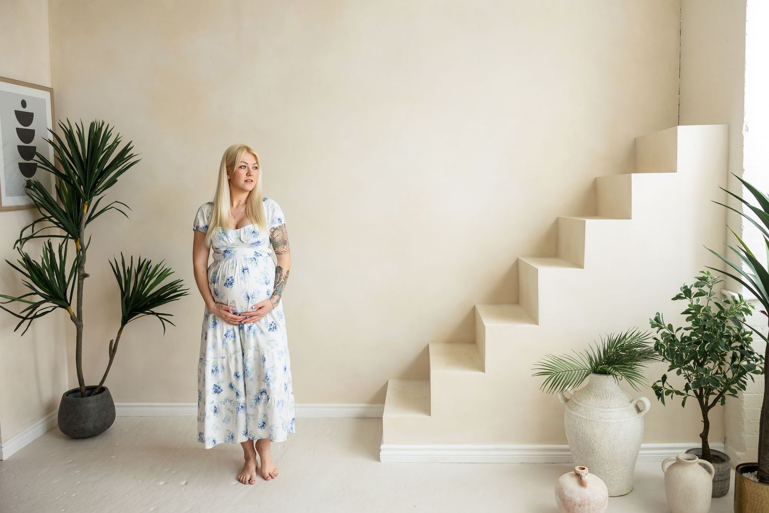 Minimalist maternity portraits against white walls with indoor plants and stairs.
