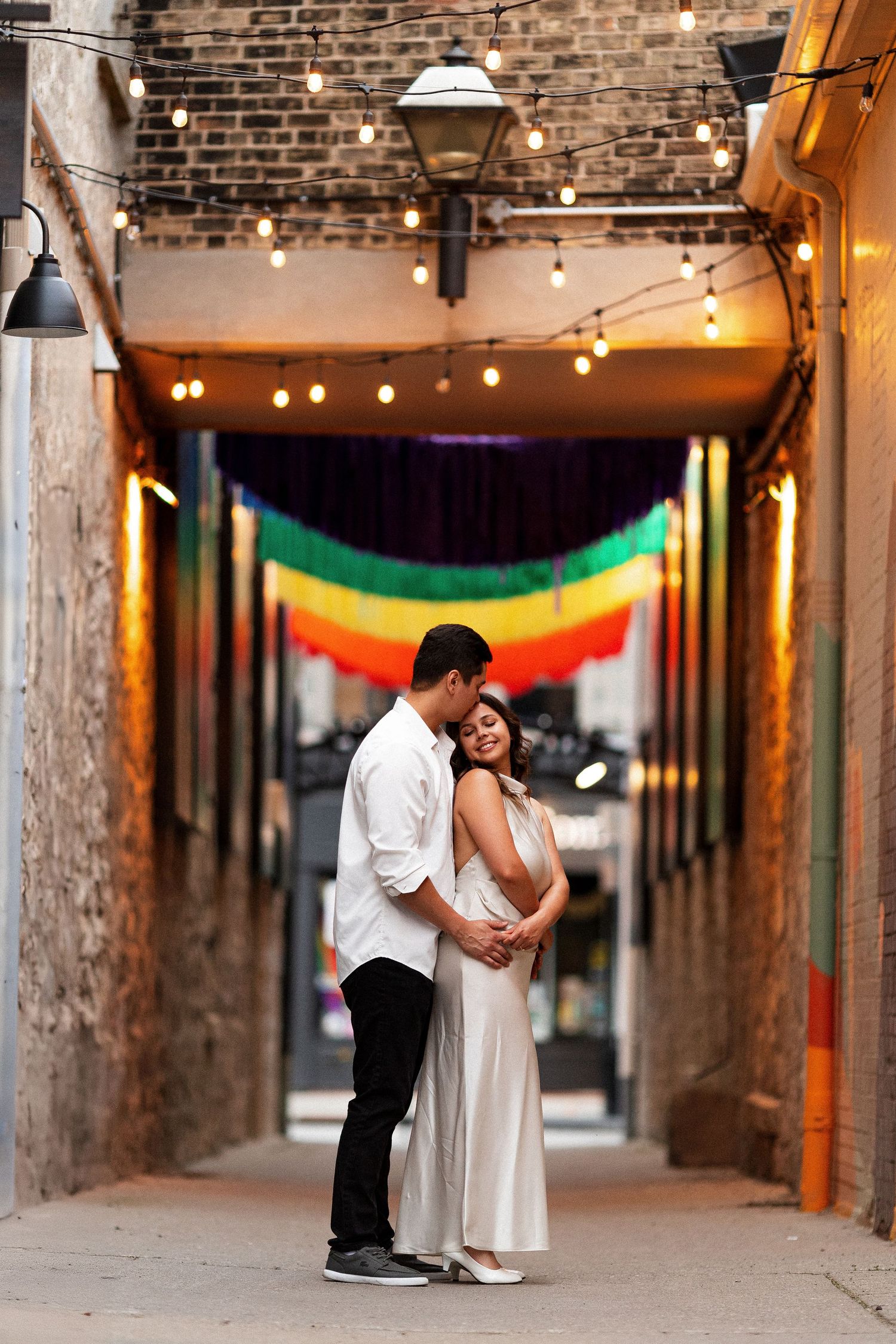 Couple embracing in a warmly lit alley decorated with string lights and a rainbow flag backdrop.