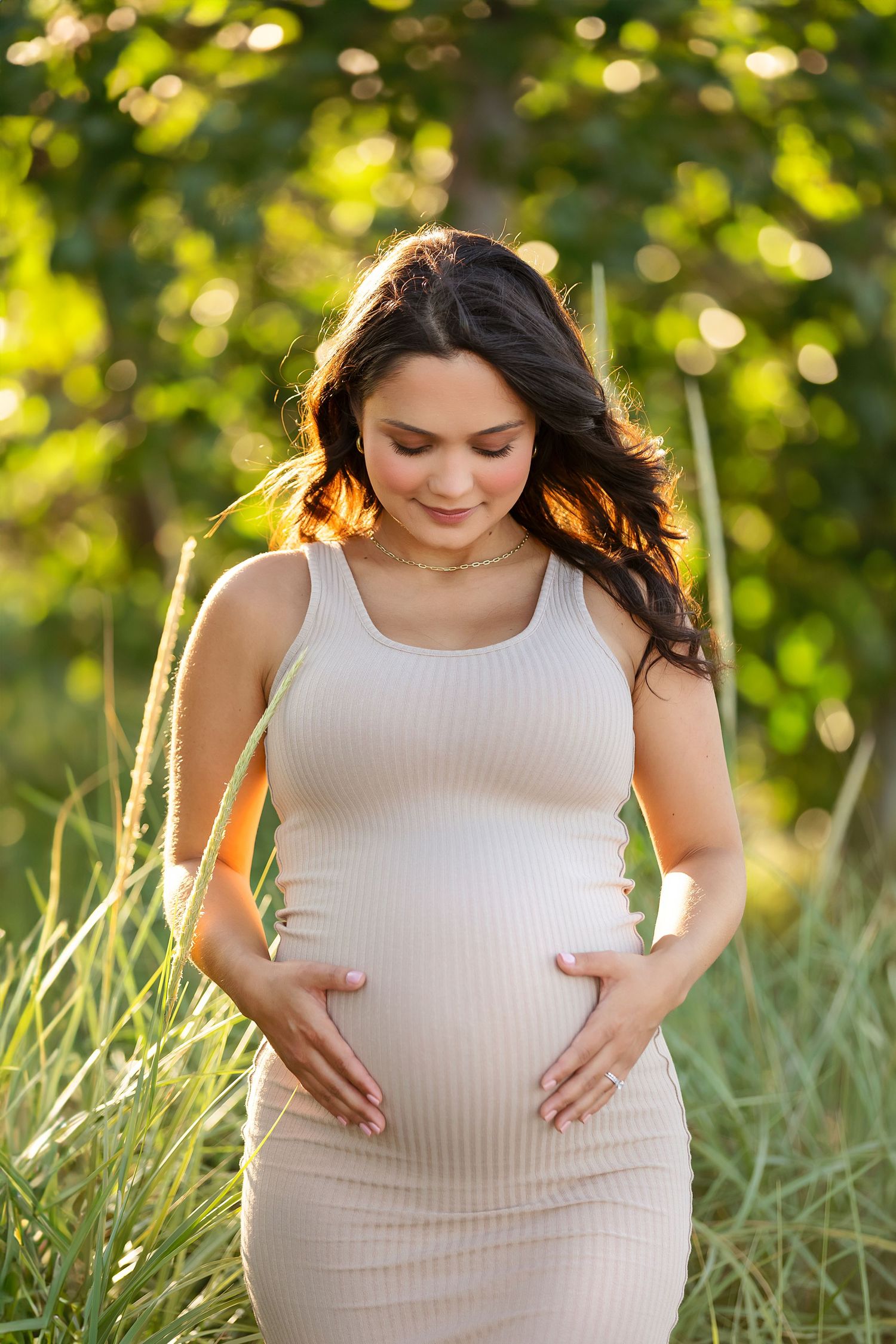 Expectant mother in cream dress poses in sunlit field with natural bokeh background.