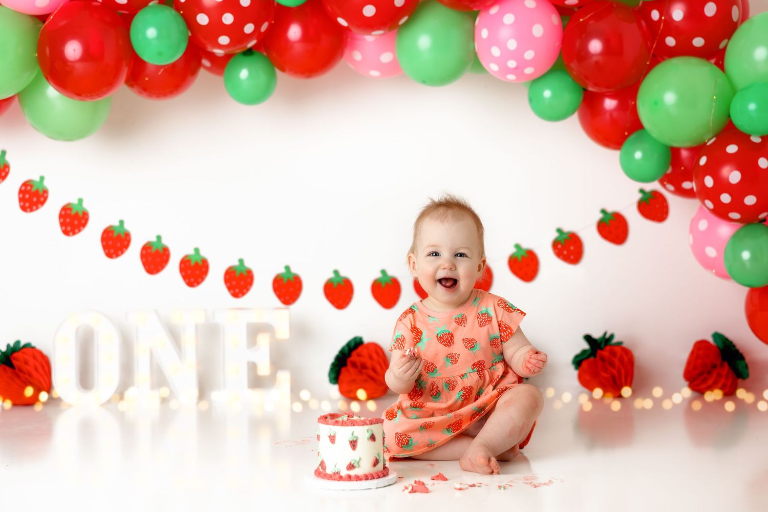 First birthday setup with red and green balloons, strawberry garland, and pink smash cake.
