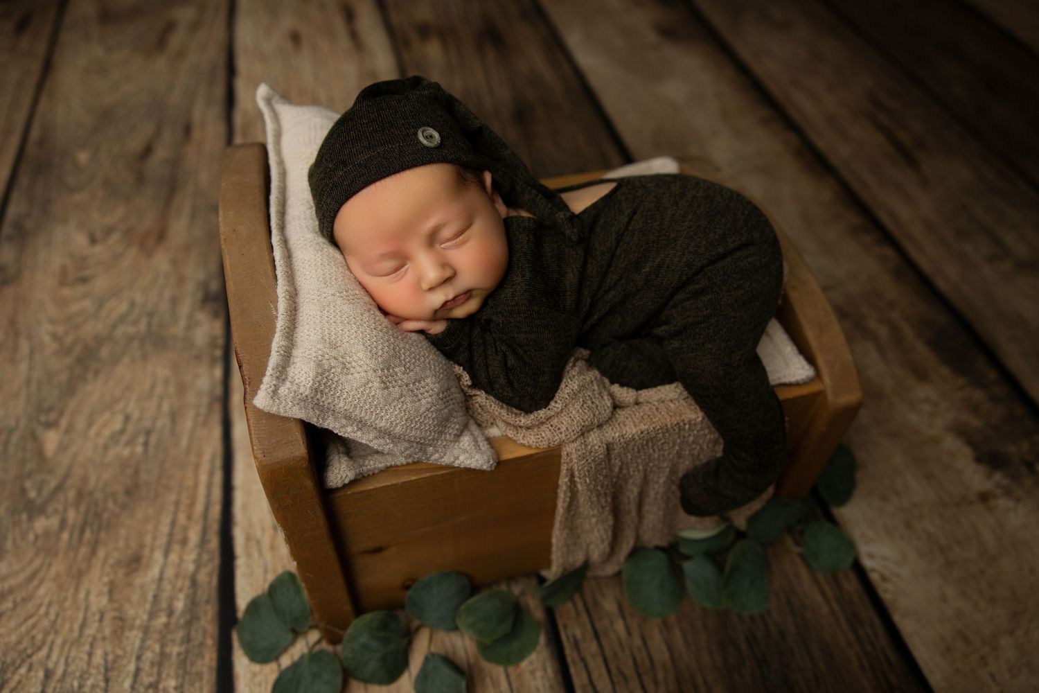 Sleeping newborn in black outfit nestled in wooden bed on rustic floor.