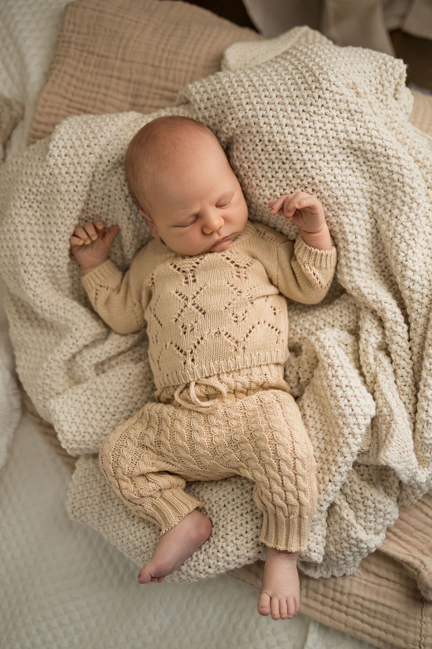 A sleeping baby wearing a cozy beige knit sweater and pants set rests peacefully on a cream-colored blanket.