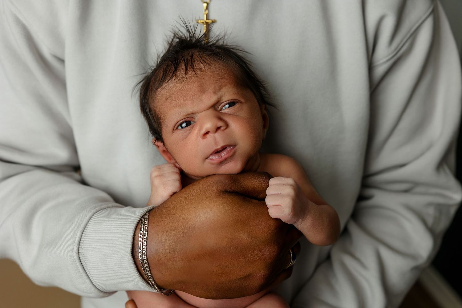 A newborn baby gazes directly at the camera while being cradled in an adult's arms wearing a gray sweatshirt.