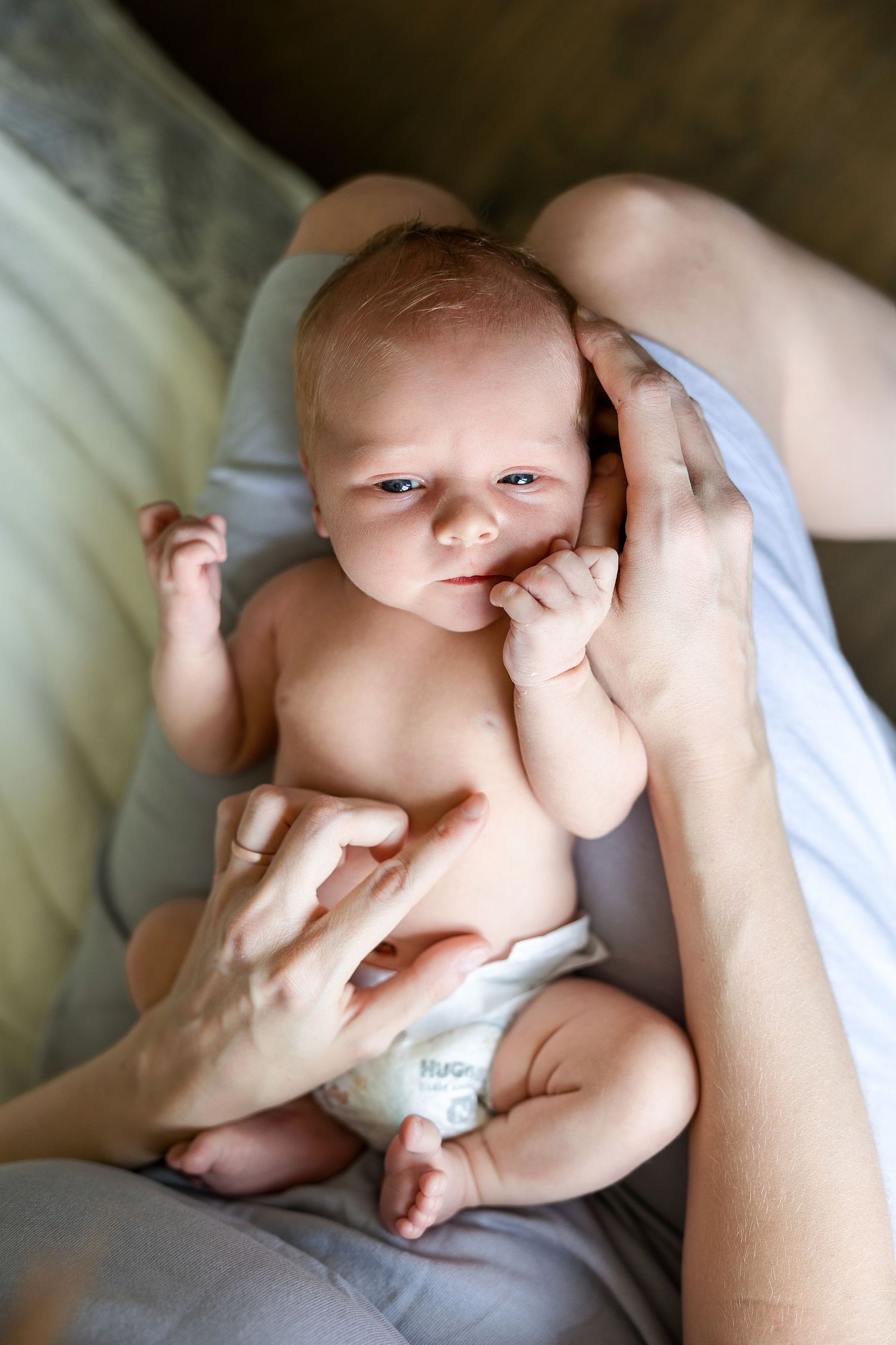 A tiny baby wearing a diaper lies on soft fabric while being caressed by gentle hands.