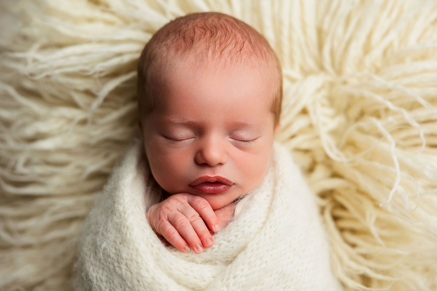Peaceful sleeping newborn wrapped in white blanket on textured cream backdrop.