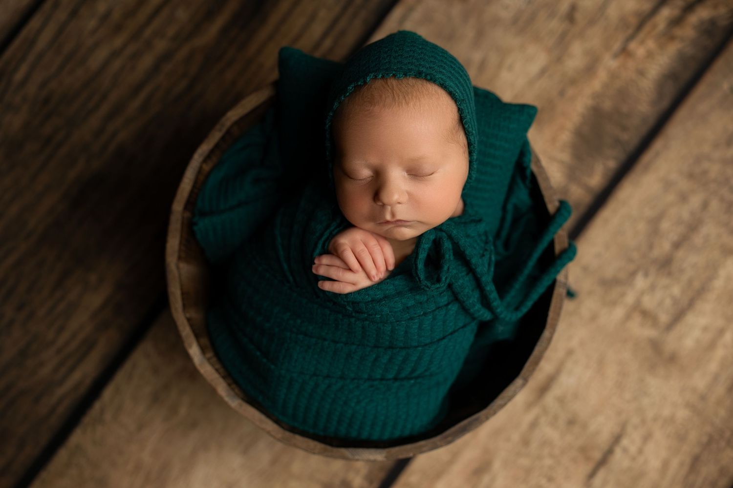Sleeping newborn wrapped in emerald green knit blanket in wooden bowl on rustic floor.