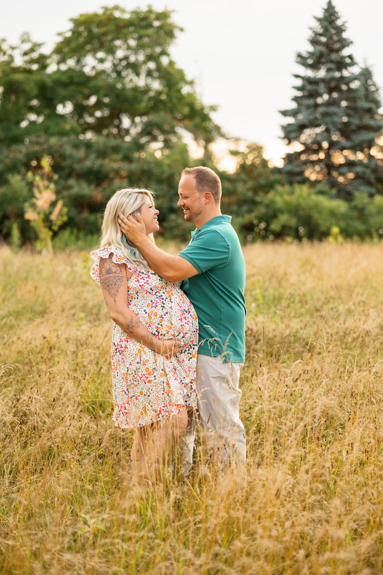 Couple dancing together in golden field at sunset with evergreen trees in background.