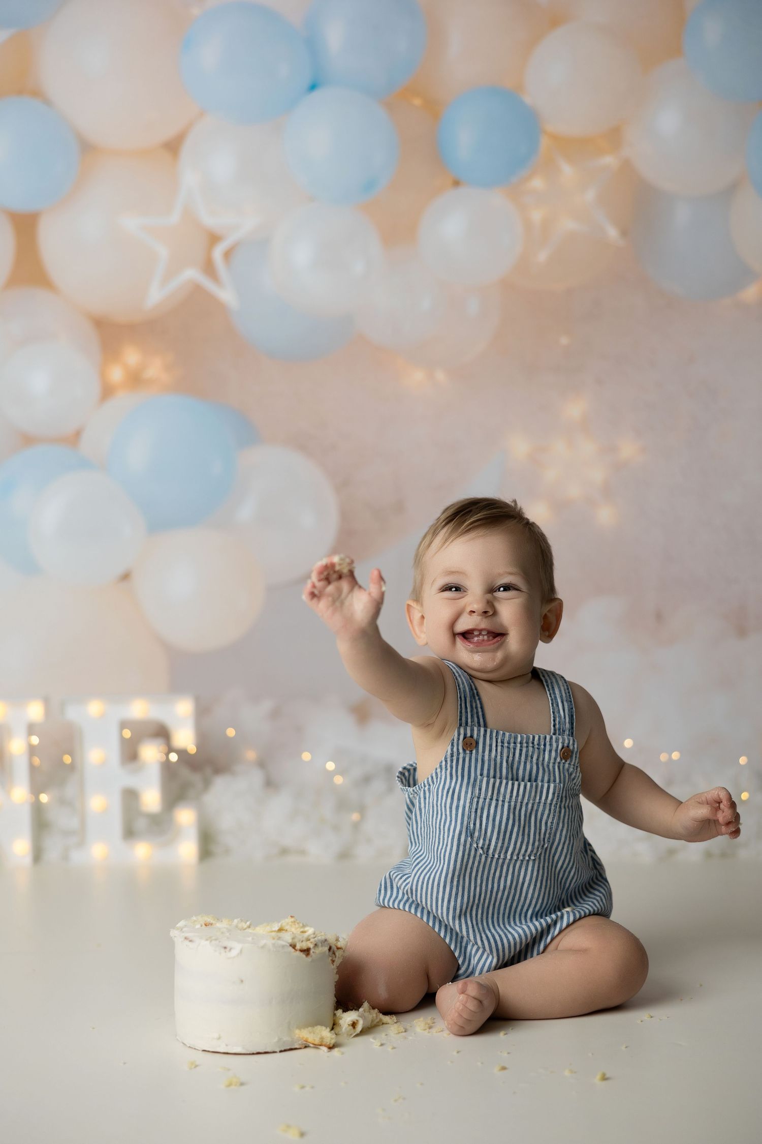 Baby in blue striped romper sits with white cake against balloon backdrop with twinkle lights.