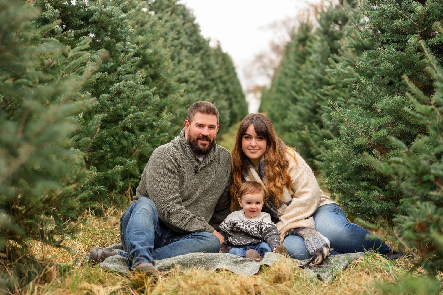 Family sitting together on ground at Christmas tree farm surrounded by evergreens.