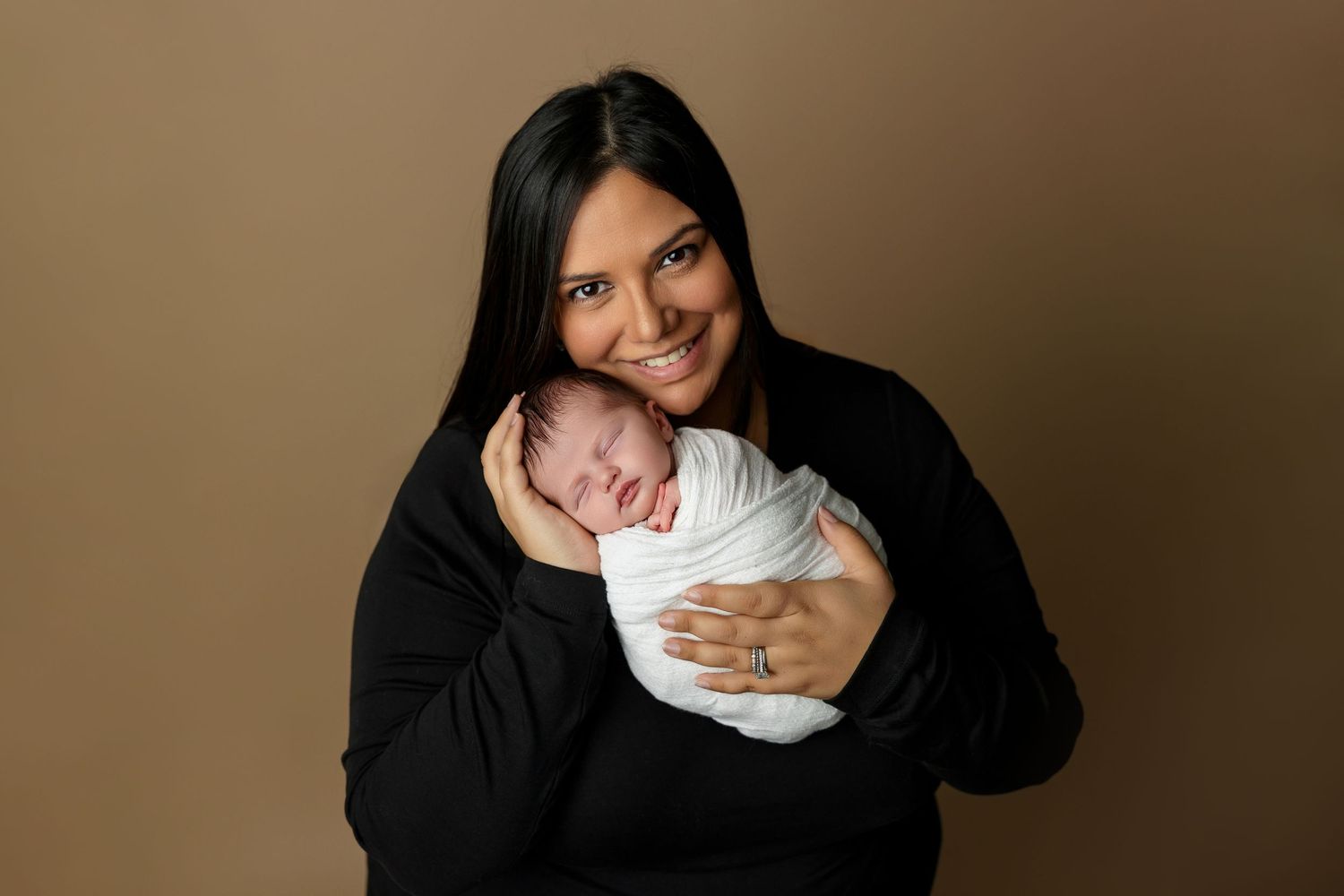 Woman in black sweater holding peaceful sleeping newborn against warm brown backdrop.