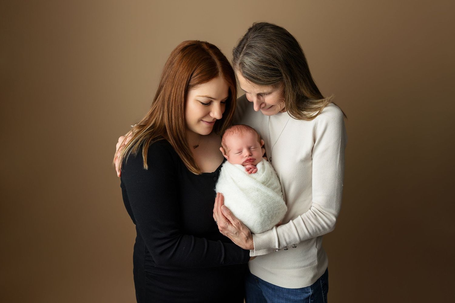 Two people share intimate moment with swaddled newborn against brown backdrop.