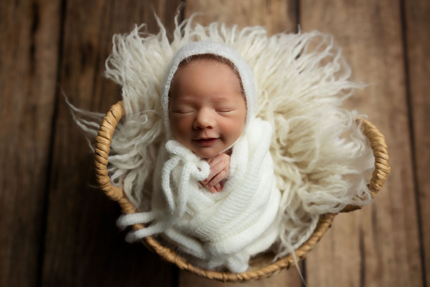 Smiling newborn wrapped in white swaddle on fluffy white backdrop.