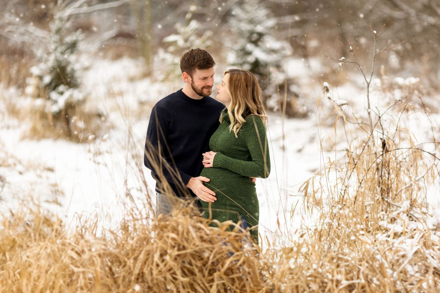 Couple embracing in winter field with tall grasses and snow in background.