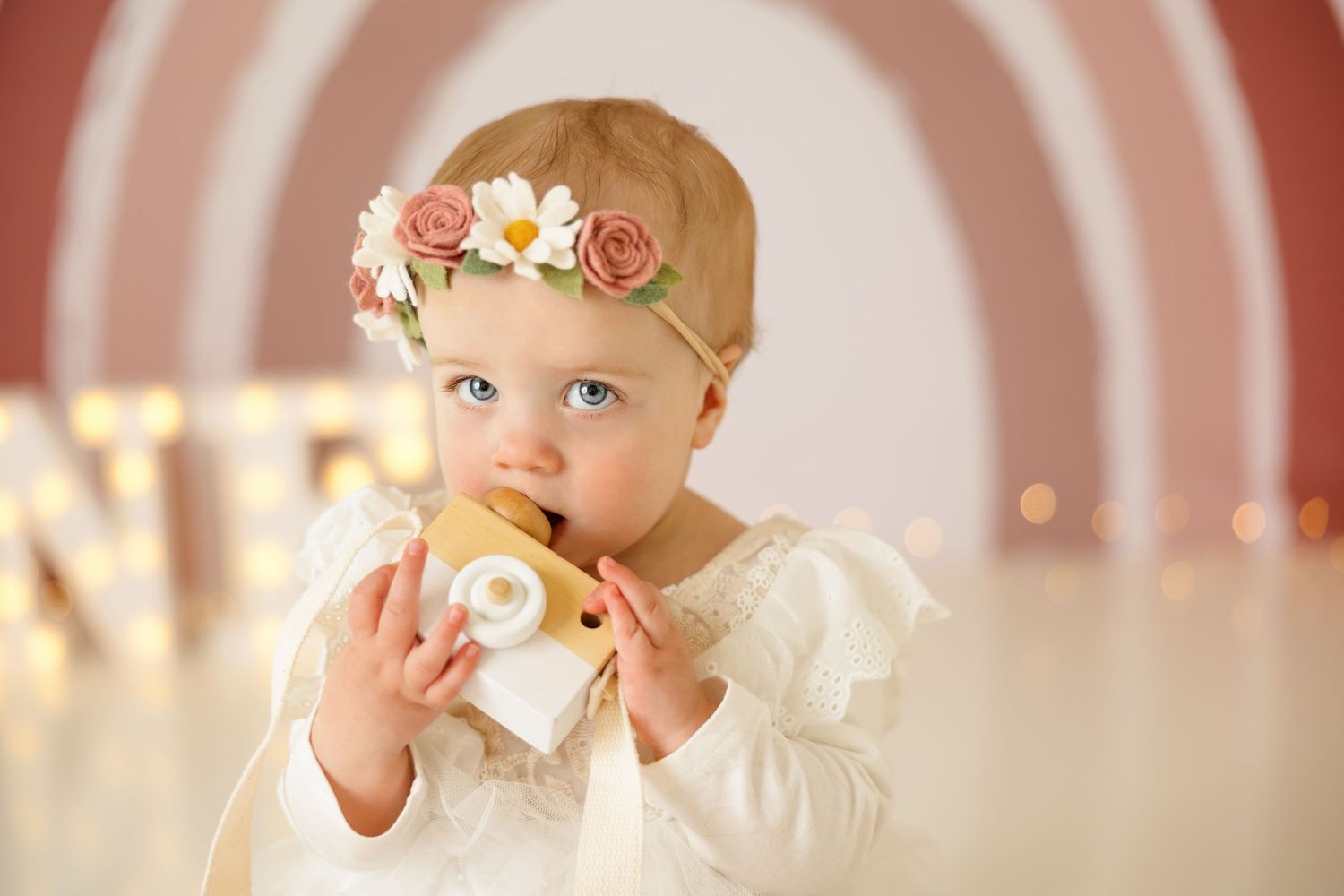 Baby with floral headband holding toy against pink circular backdrop.