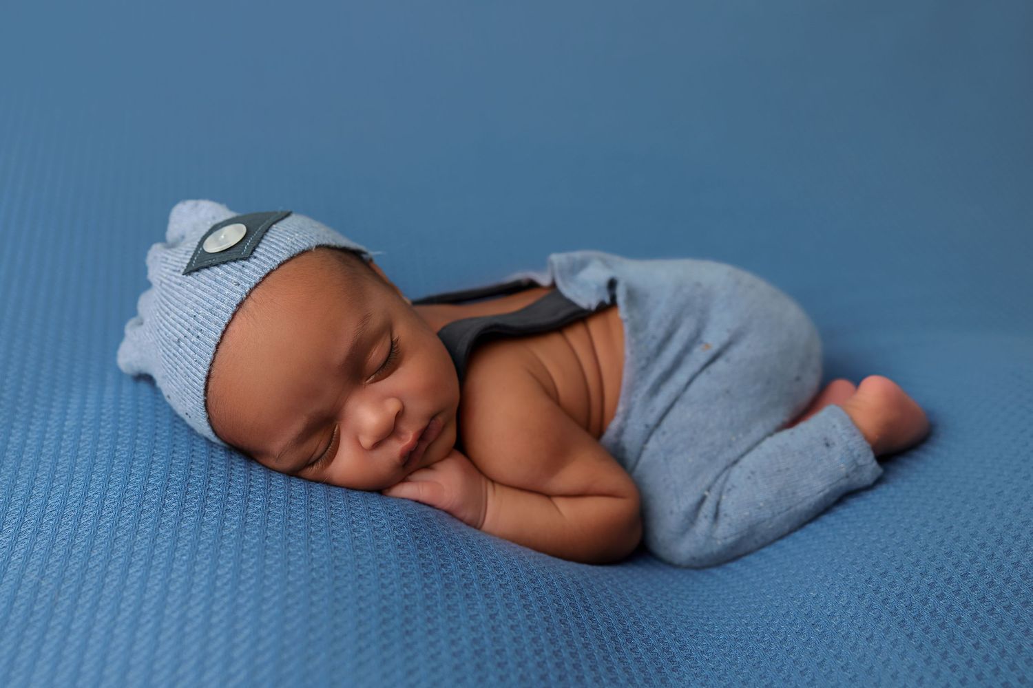 Newborn baby wearing soft blue outfit and hat sleeping peacefully on blue textured surface.