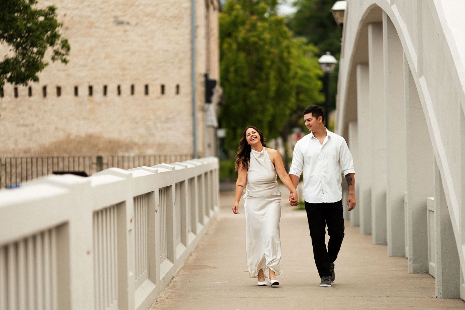A smiling couple walks hand-in-hand across a white bridge, dressed in elegant white and black outfits.