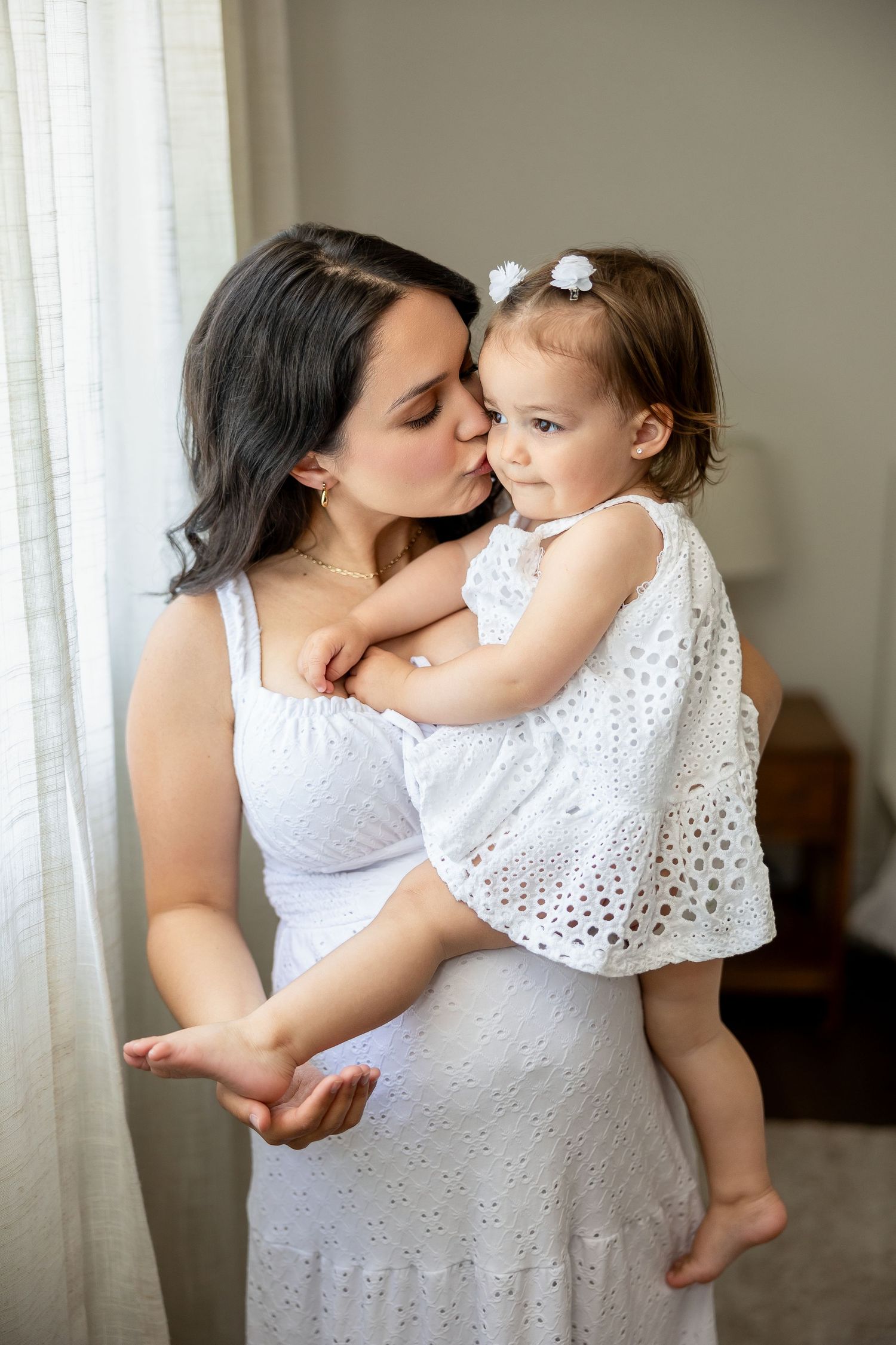 A tender moment between parent and child in matching white eyelet dresses near a window at home.