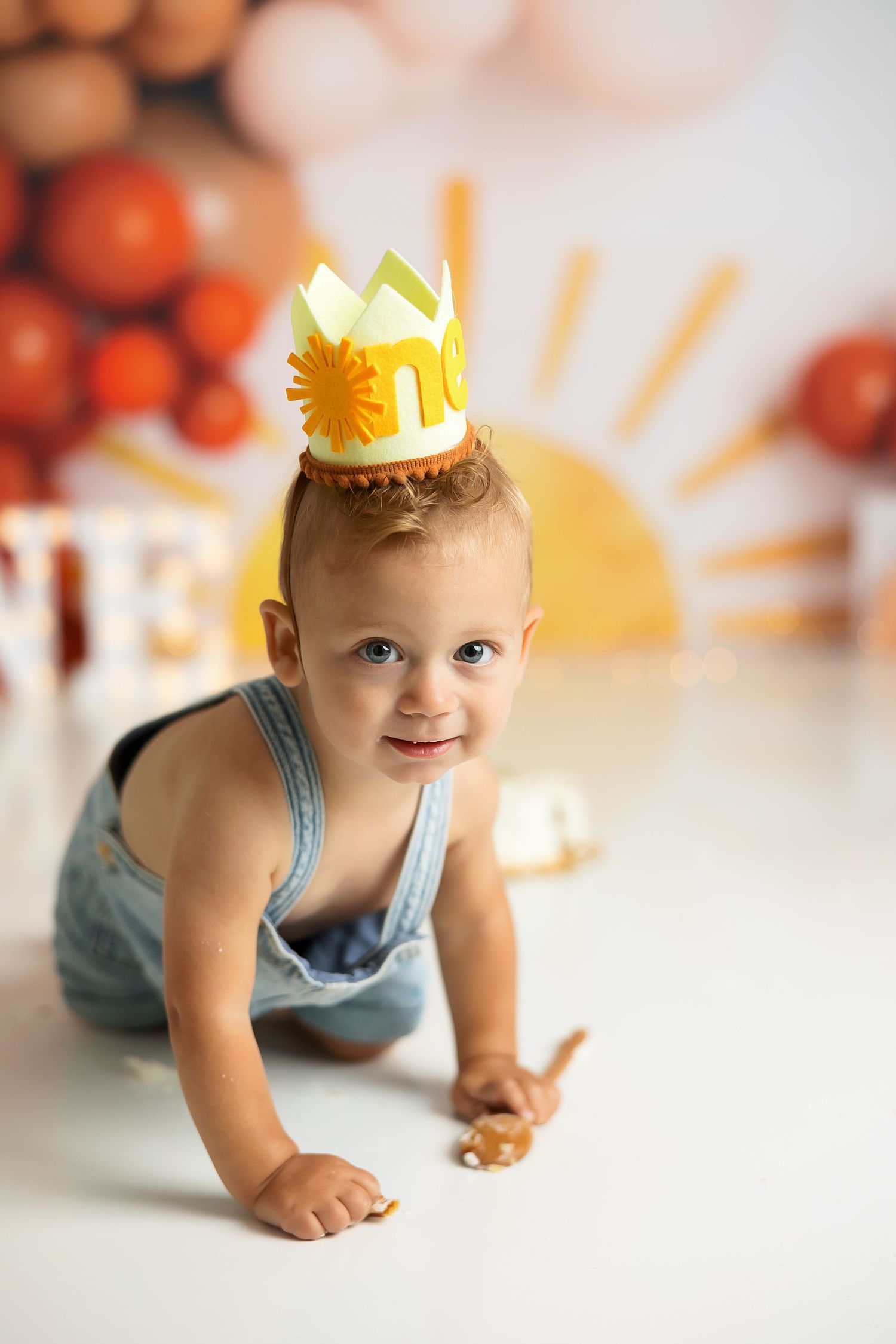 A special first birthday photo with light blue overalls and a decorative paper crown against a balloon backdrop.