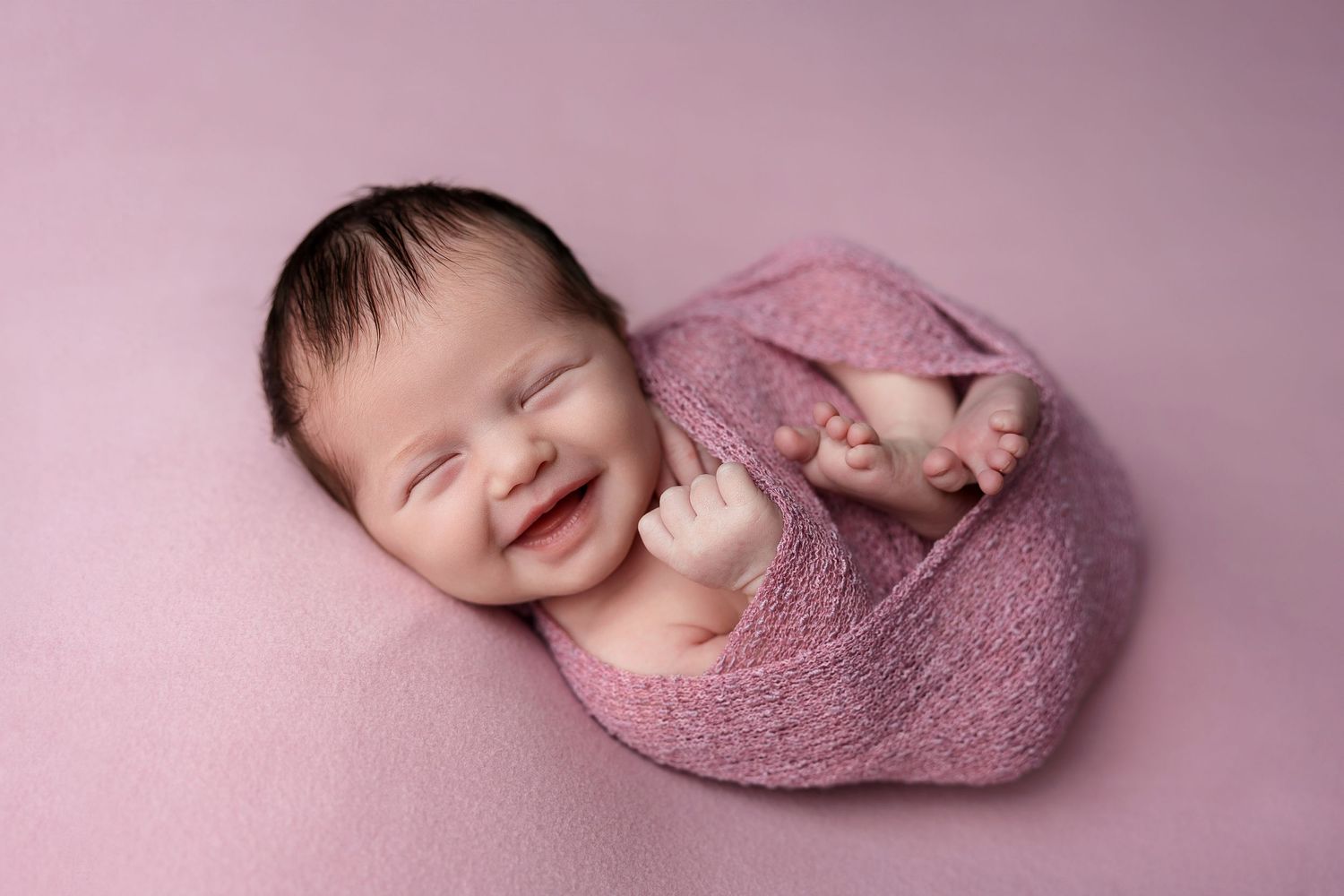 A happy newborn wrapped in pink fabric shows a bright smile against a rose-colored background.