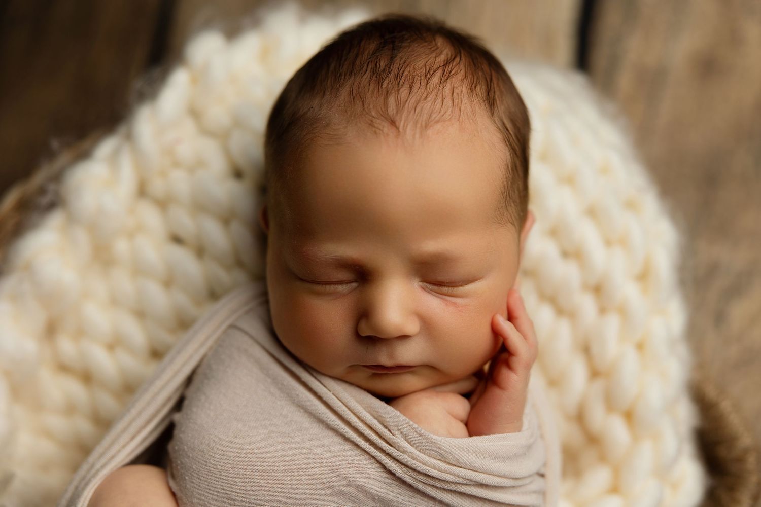 Peaceful sleeping newborn wrapped in beige swaddle on cream knit blanket.