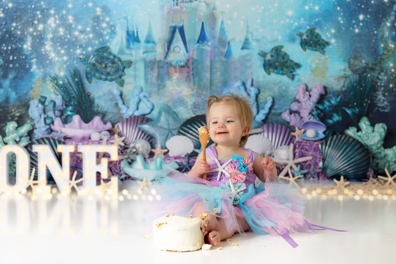 Magical first birthday photo with blue and purple underwater themed backdrop featuring lit up ONE sign.