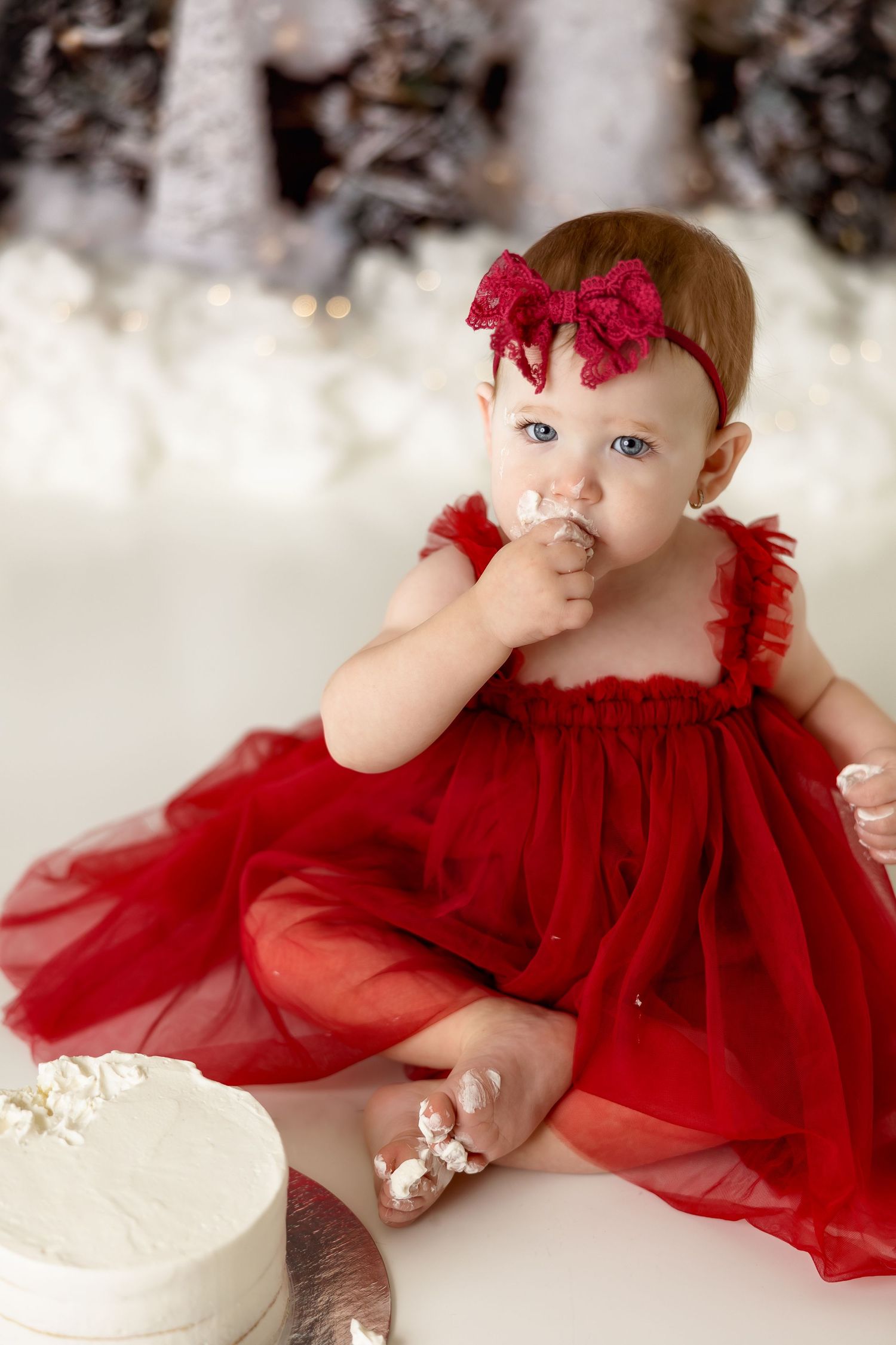 A special occasion photo of a baby in a red tulle dress with matching bow sitting next to a white cake.