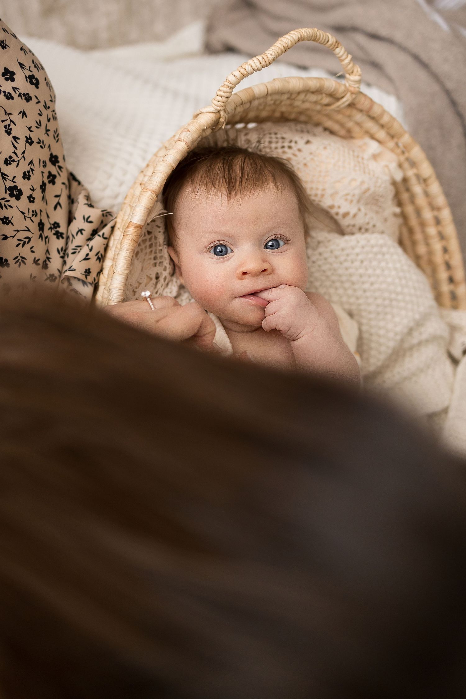 Adorable blue-eyed baby lying in a wicker Moses basket with a knit blanket, gazing up curiously.