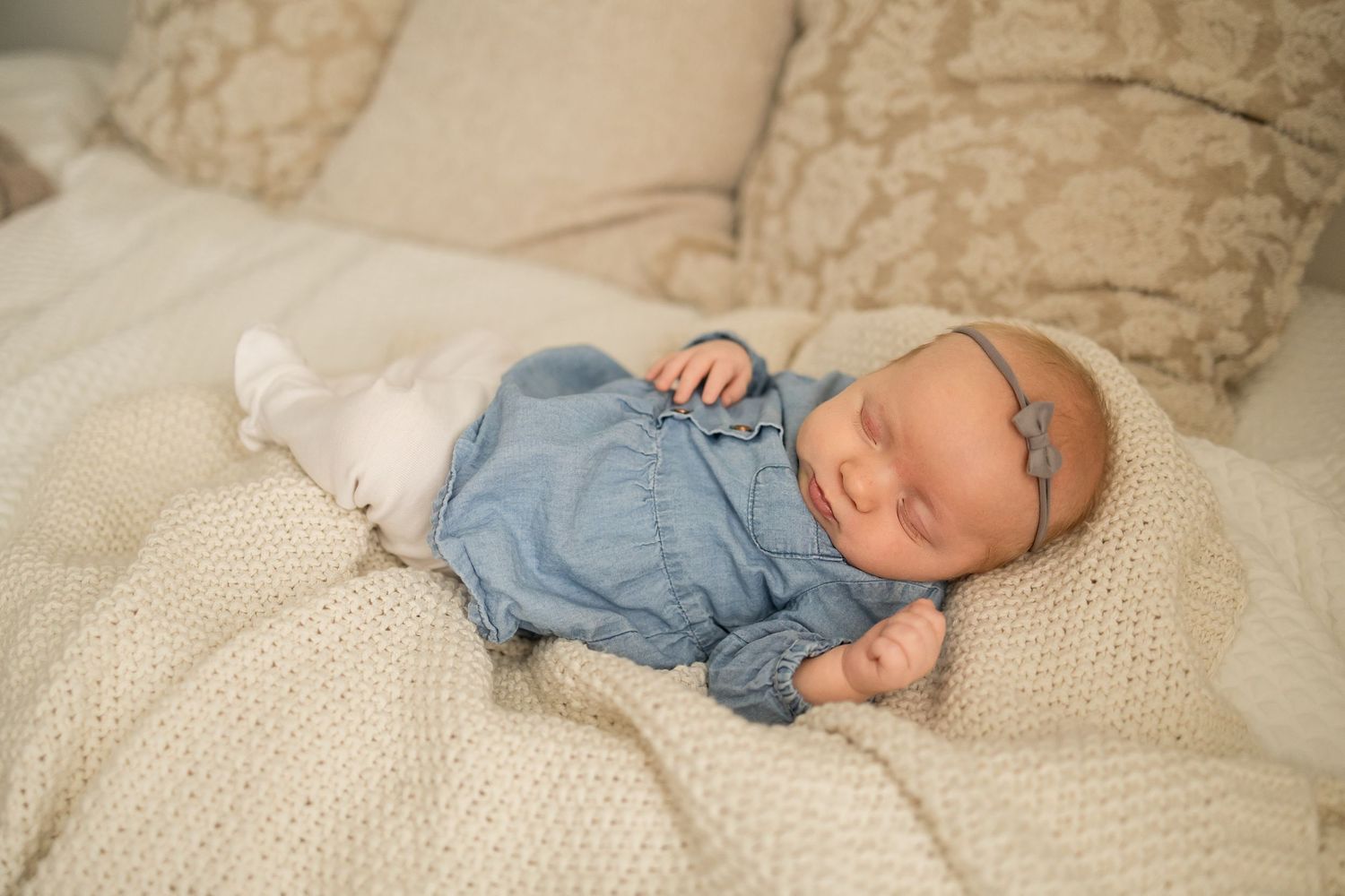 A sleeping baby wearing denim overalls rests peacefully on a cream-colored knit blanket.