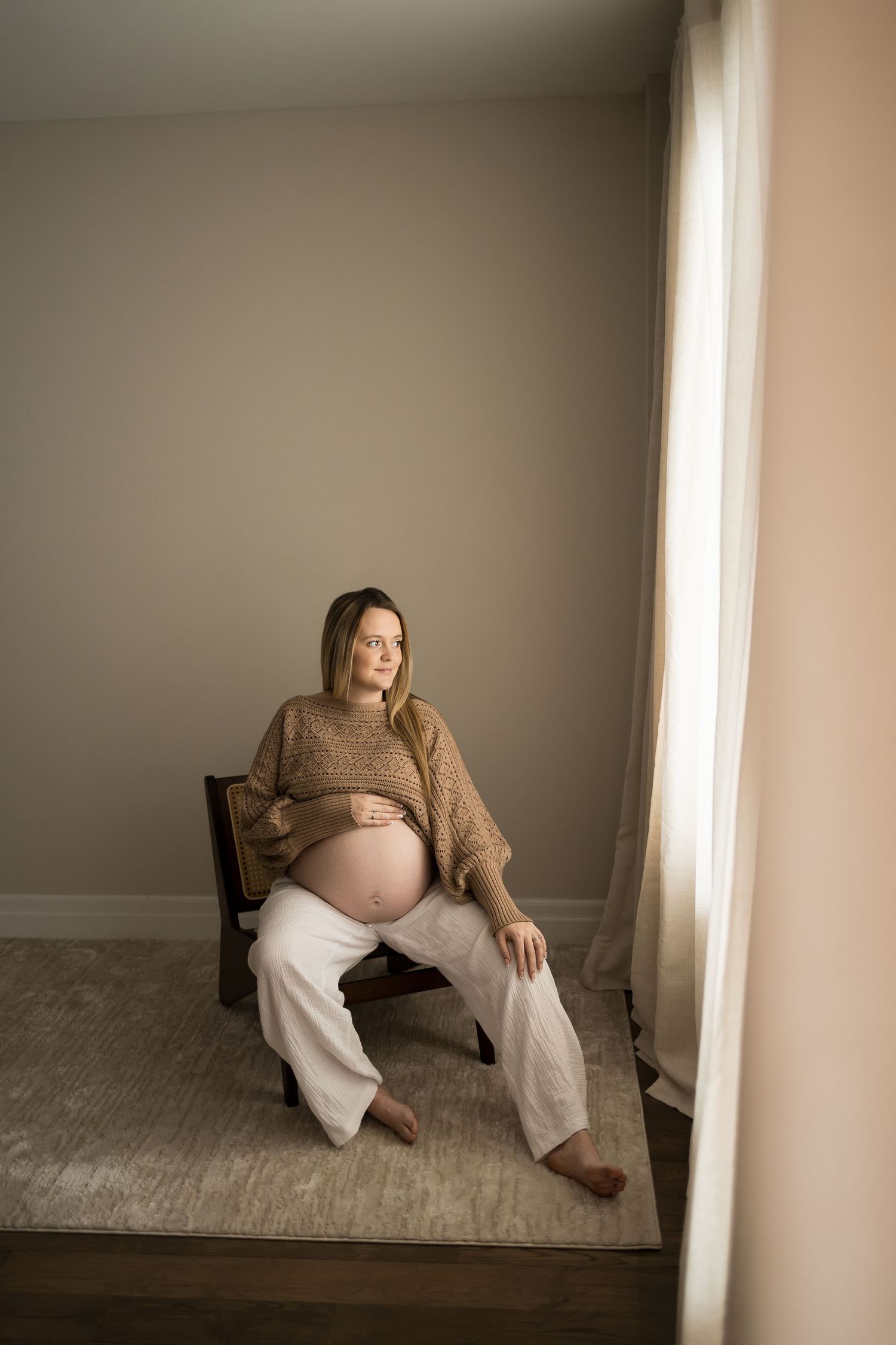A person in brown clothing sits against a wall in a minimalist room with beige tones and carpeted floor.