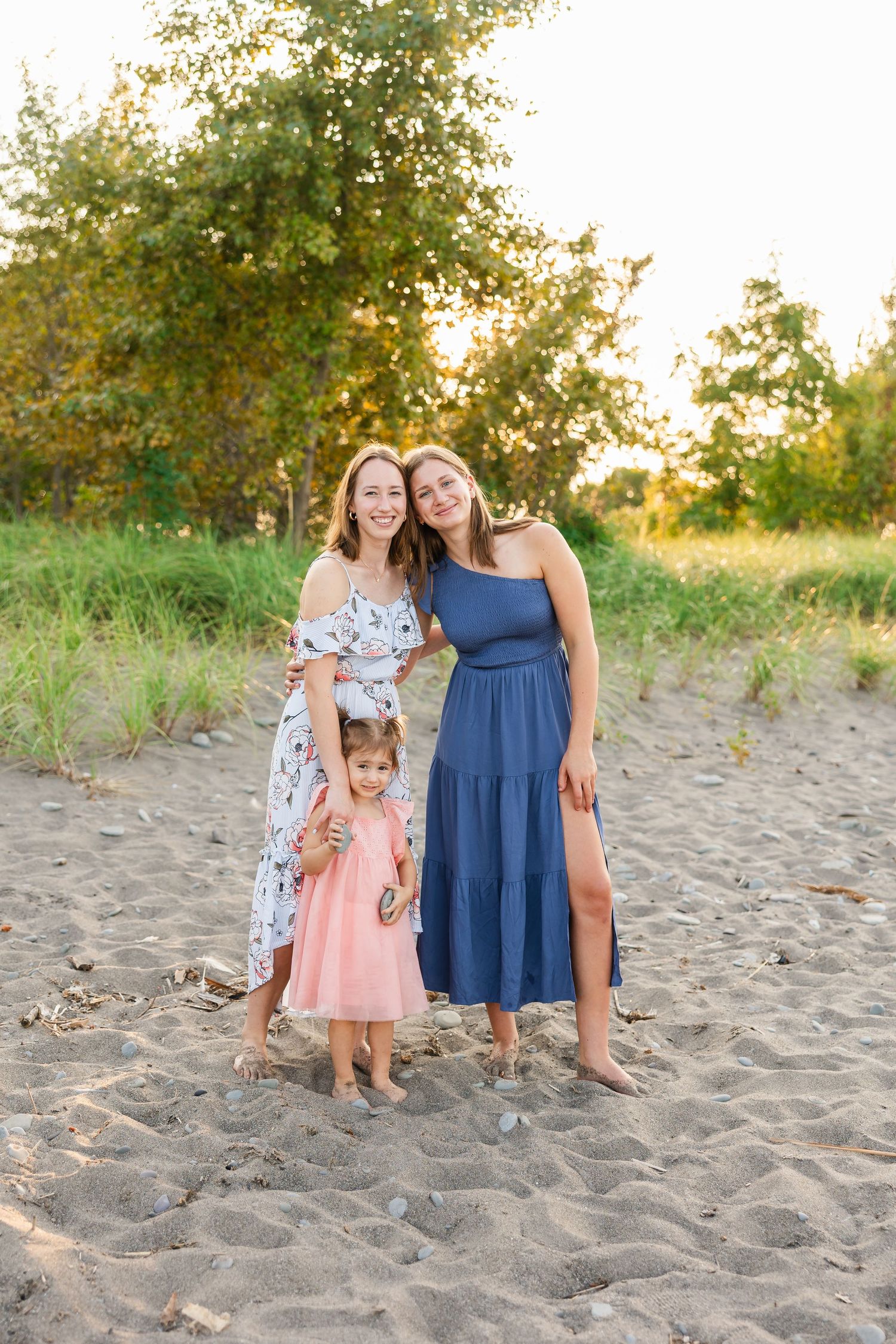 Three people wearing summer dresses pose together on a sandy beach during golden hour sunset.
