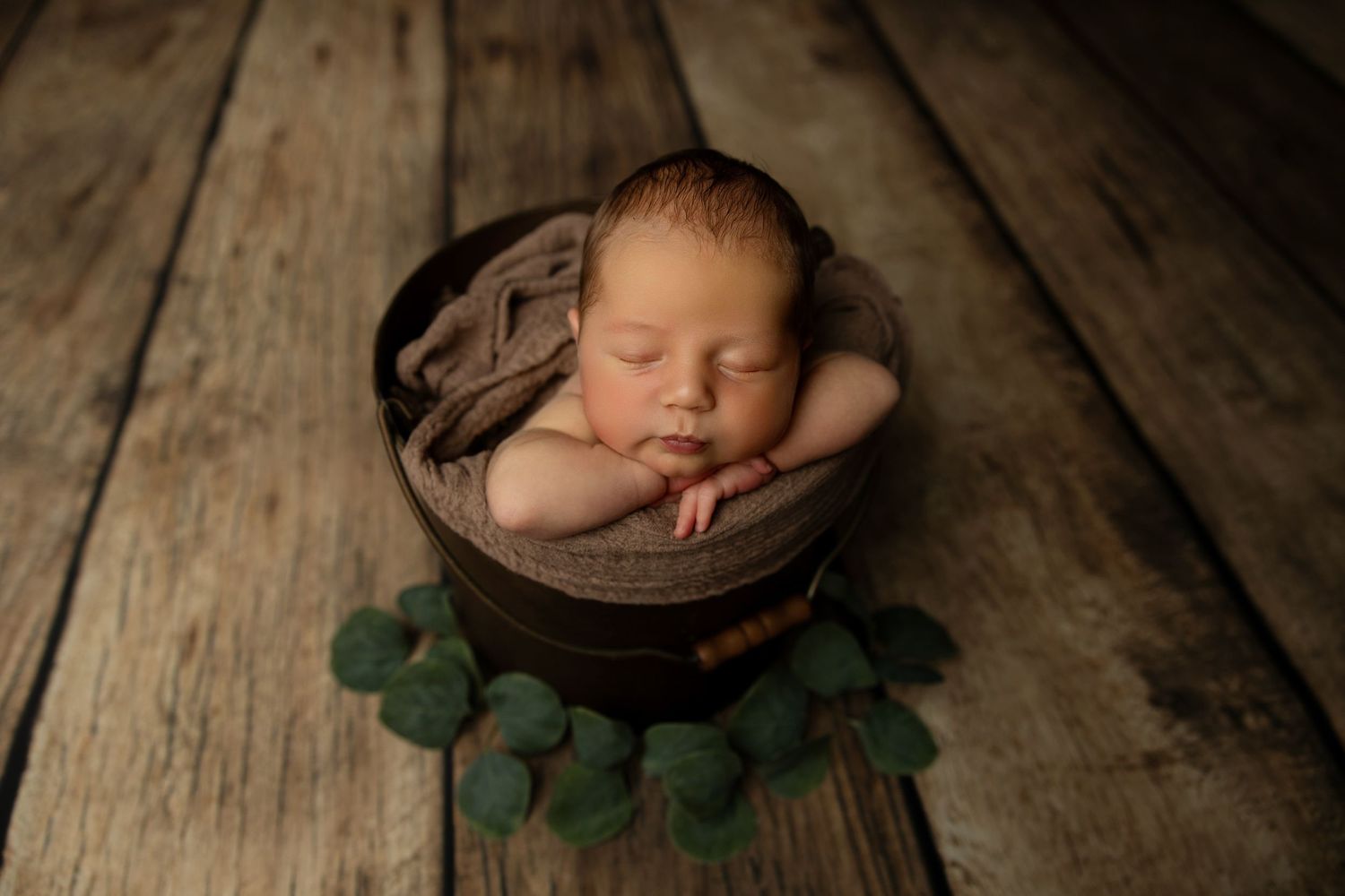 Sleeping newborn in brown bowl surrounded by green eucalyptus leaves on wooden backdrop.