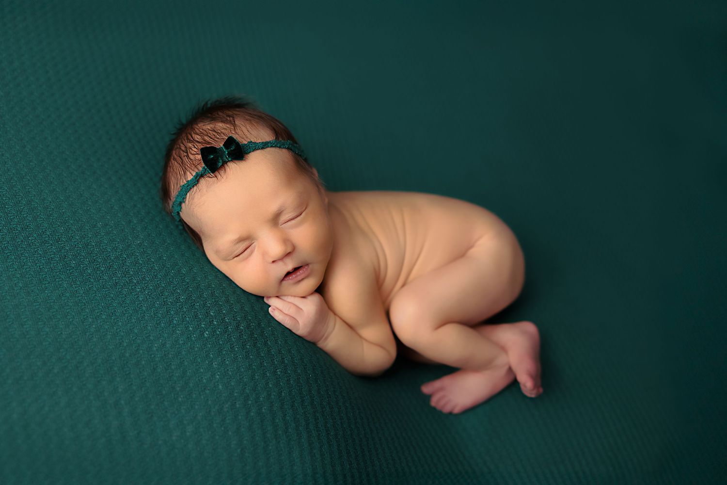 Newborn baby wearing headband posed on teal textured backdrop.
