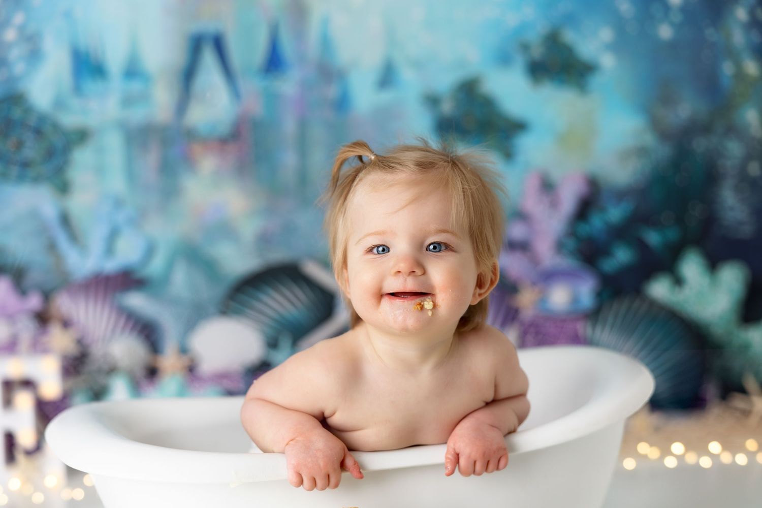 Happy baby posing in white bathtub with magical underwater themed birthday backdrop.