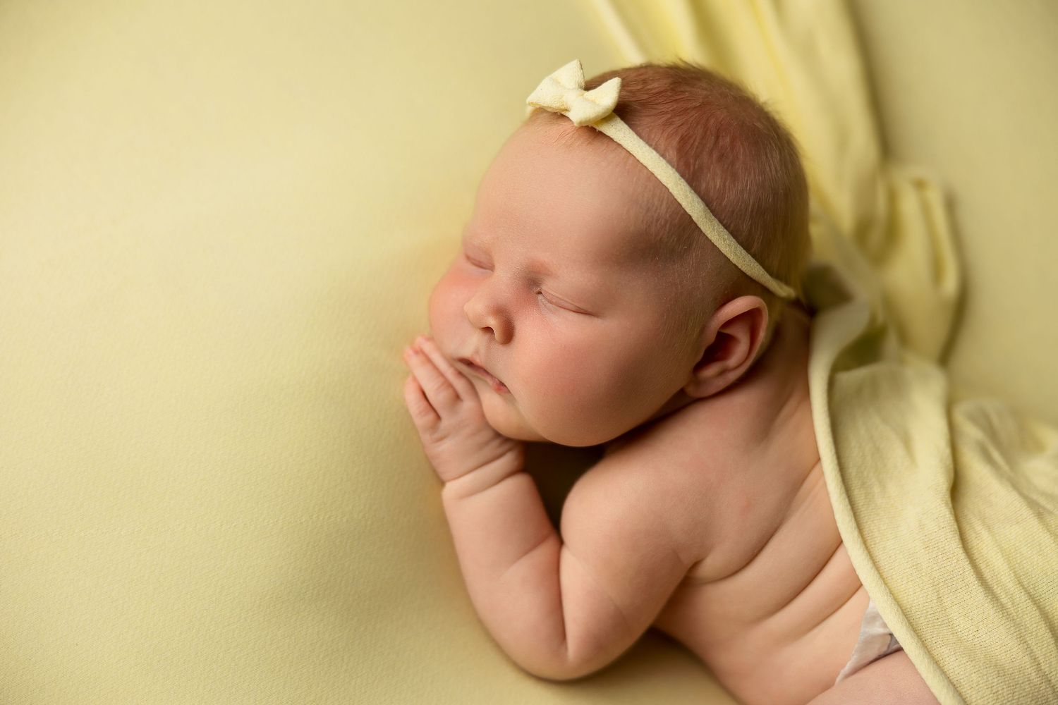 Newborn baby sleeping peacefully on yellow blanket wearing delicate bow headband.